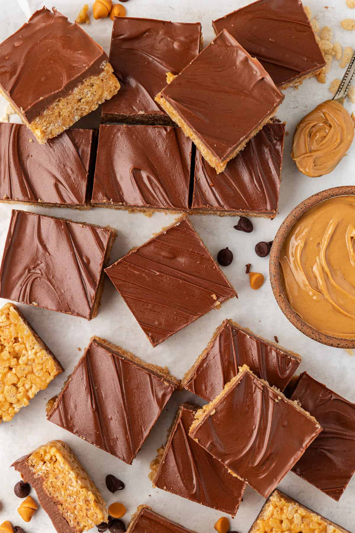 over head view of scotcheroos scattered around on a white surface with butterscotch and chocolate chips and rice cereal sprinkled around, a bowl of peanut butter and a spoonful of peanut butter