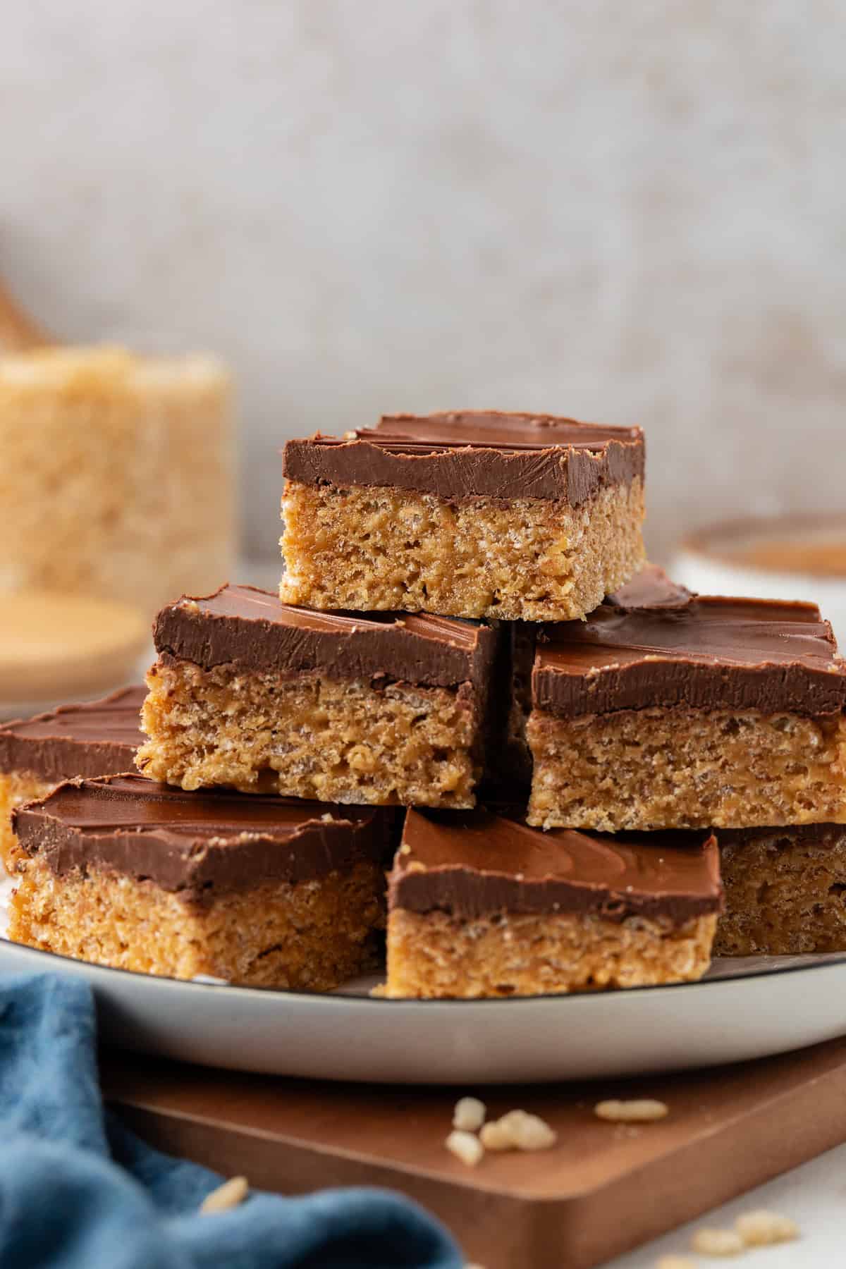 a plate full of scotcheroos stacked up in 3 layers on top of a wooden platter with a blue towel beside it and rice cereal sprinkled around