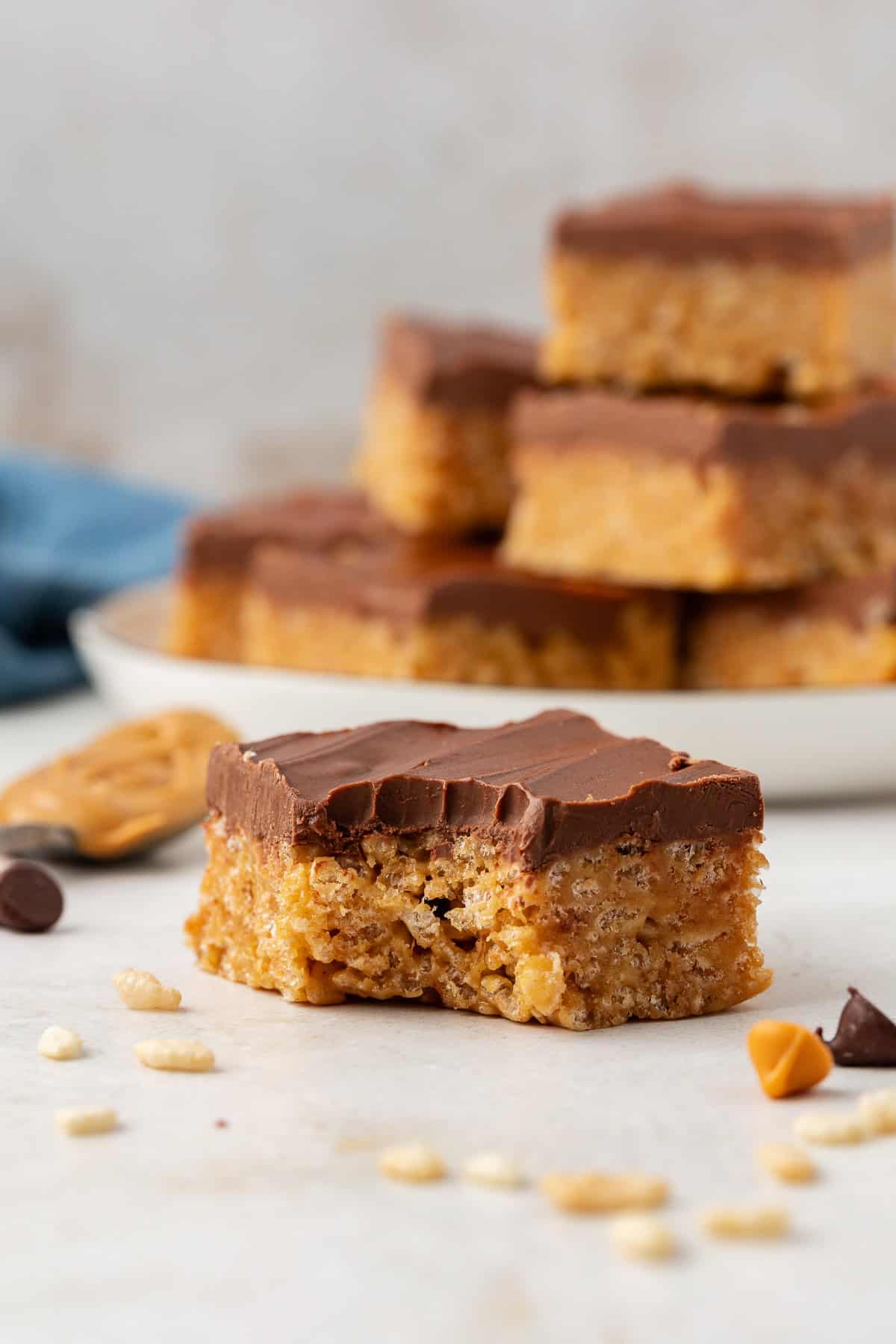 a scotcheroo on a countertop surface surrounded by rice cereal, chocolate chips and butterscotch chips scattered around, and a plate full of scotcheroos in the background