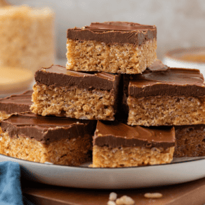 a plate full of scotcheroos stacked up in 3 layers on top of a wooden platter with a blue towel beside it and rice cereal sprinkled around