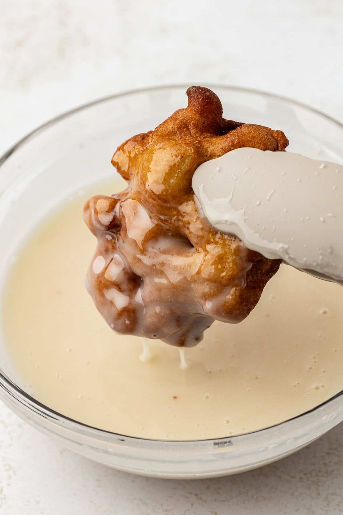 an apple fritter being dipped into a bowl of glaze with tongs