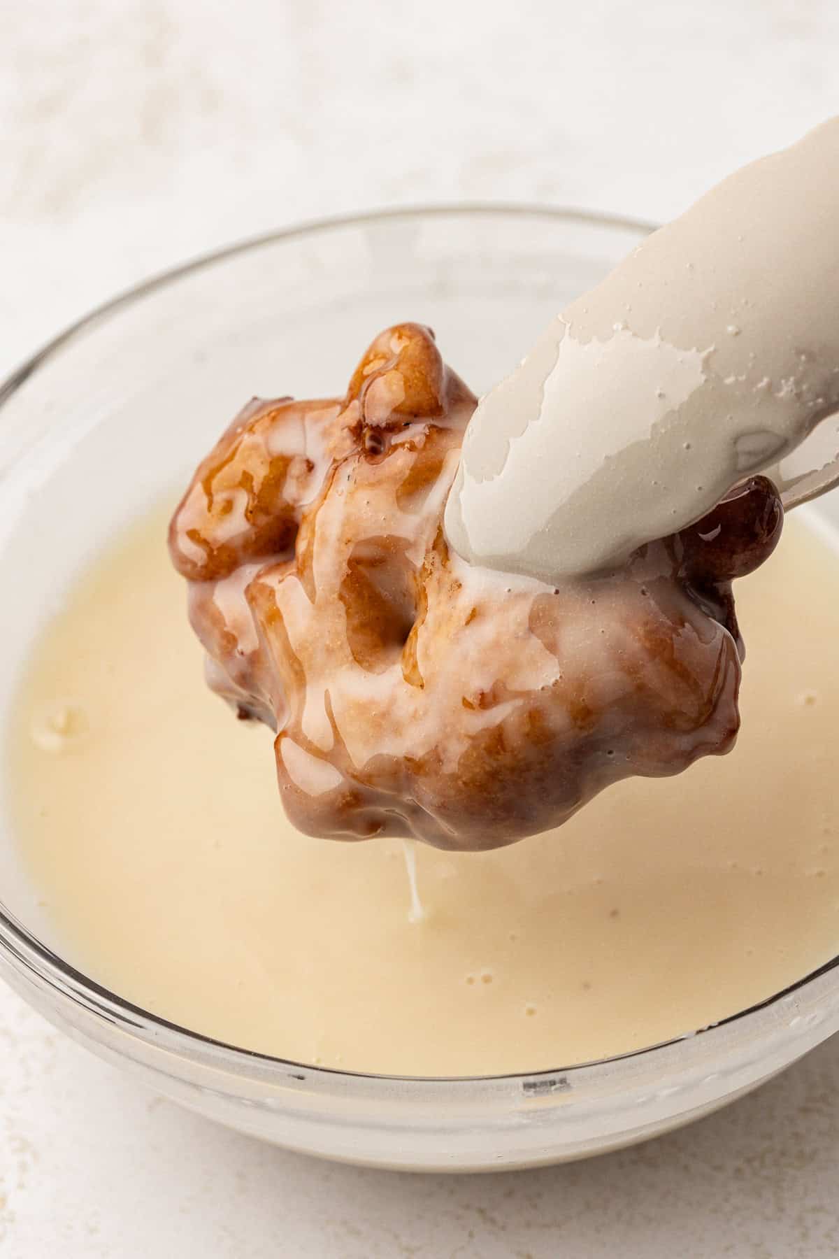 an apple fritter held over a bowl of glaze with tongs, covered in and dripping glaze back into the bowl