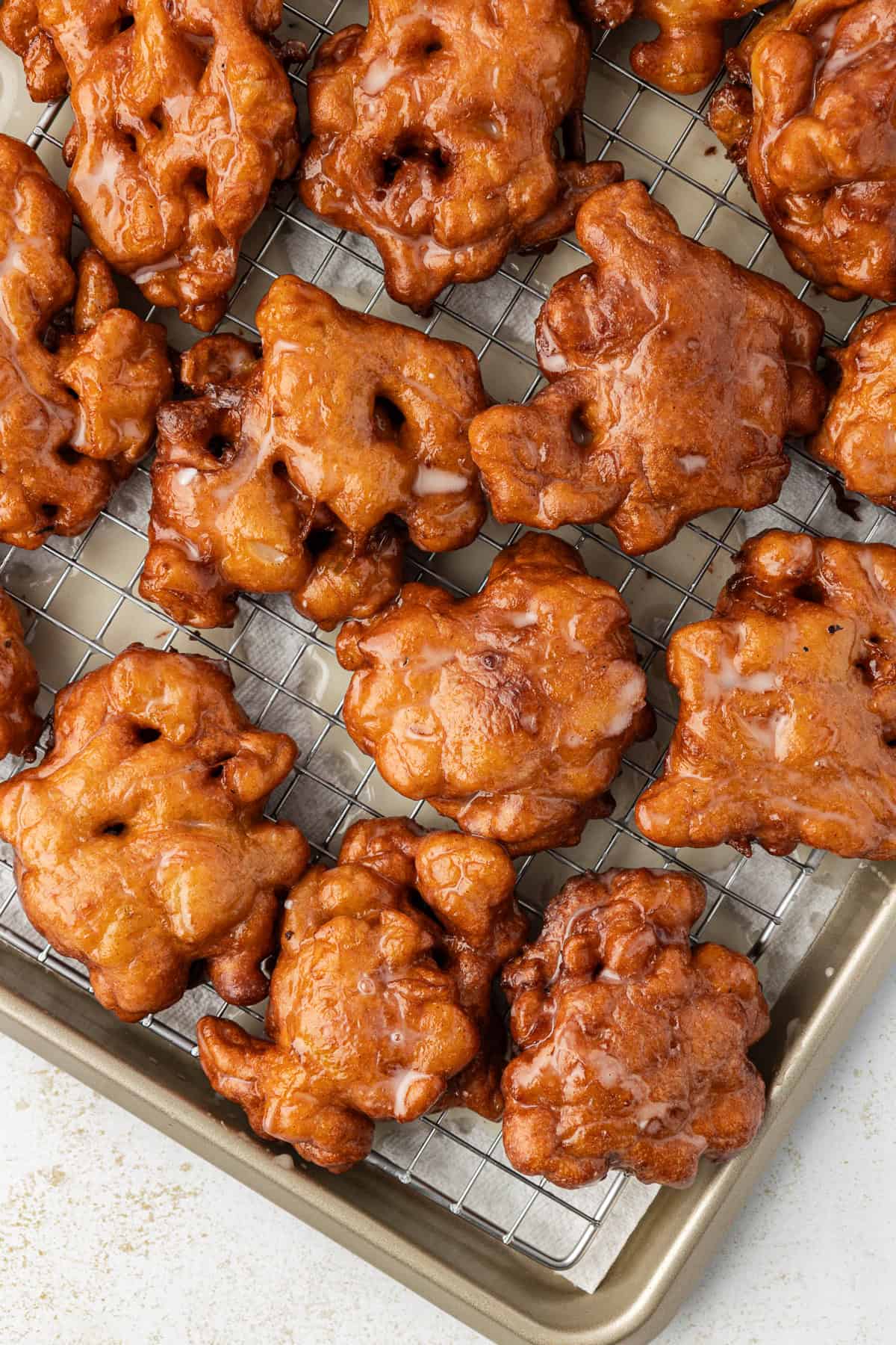 apple fritters lined in rows on a wire rack on top of a baking sheet lined with paper towels