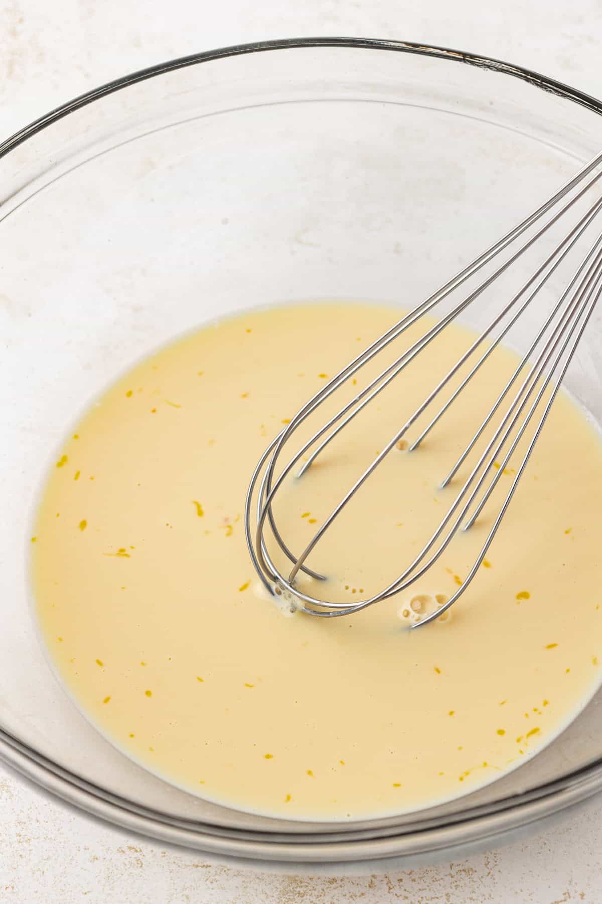 wet ingredients for apple fritters in a clear glass bowl with a whisk