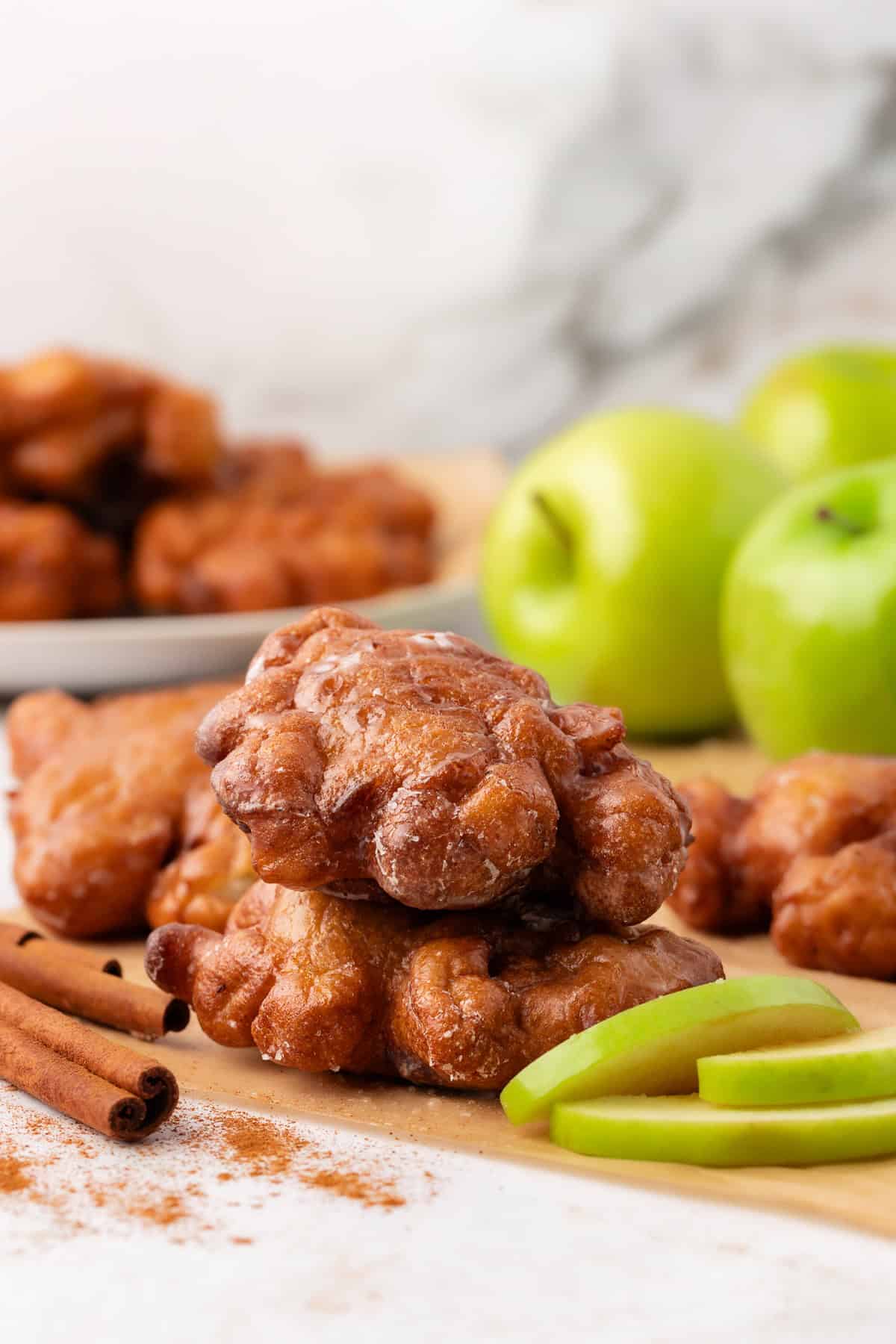 two apple fritters stacked on brown paper, surrounded by green apple slices, cinnamon sticks, and more fritters and whole green apples in the background