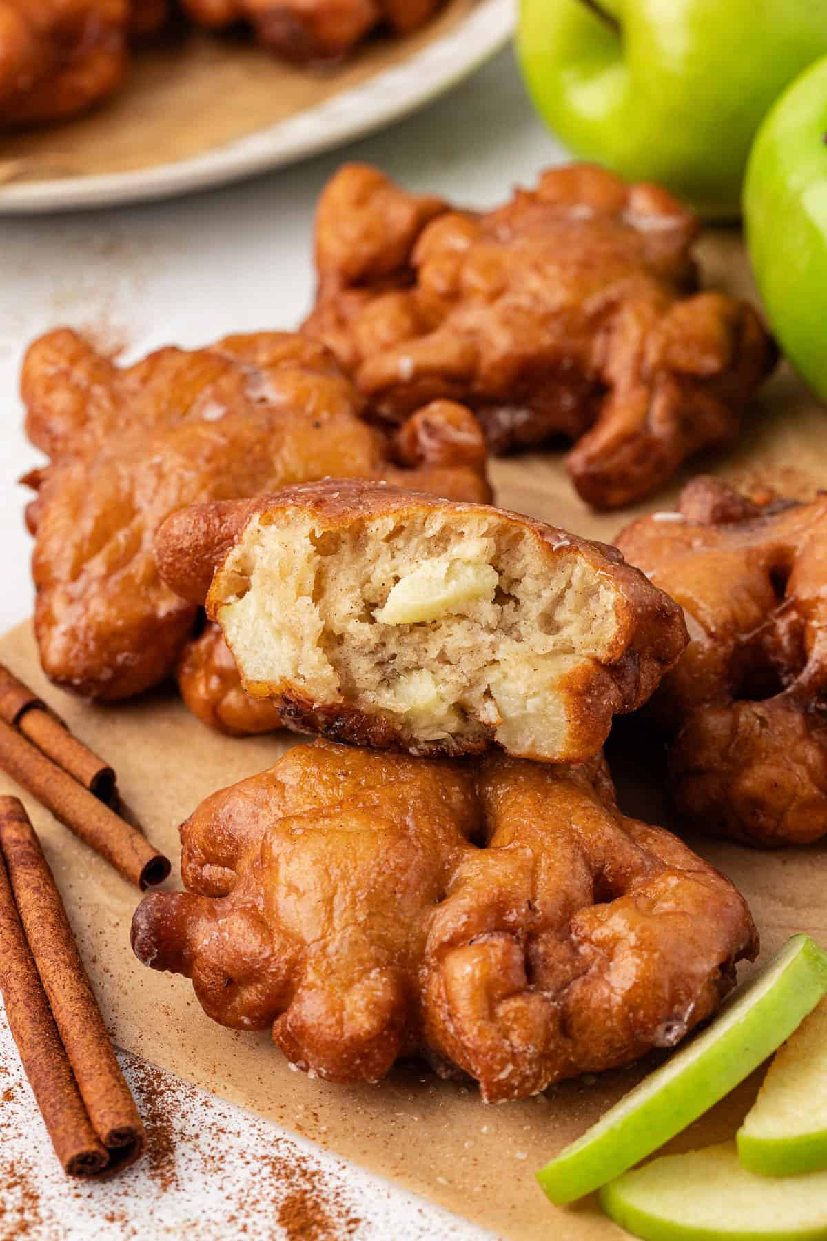 apple fritters scattered around on a piece of brown paper, surrounded by cinnamon sticks, green apples and green apple slices