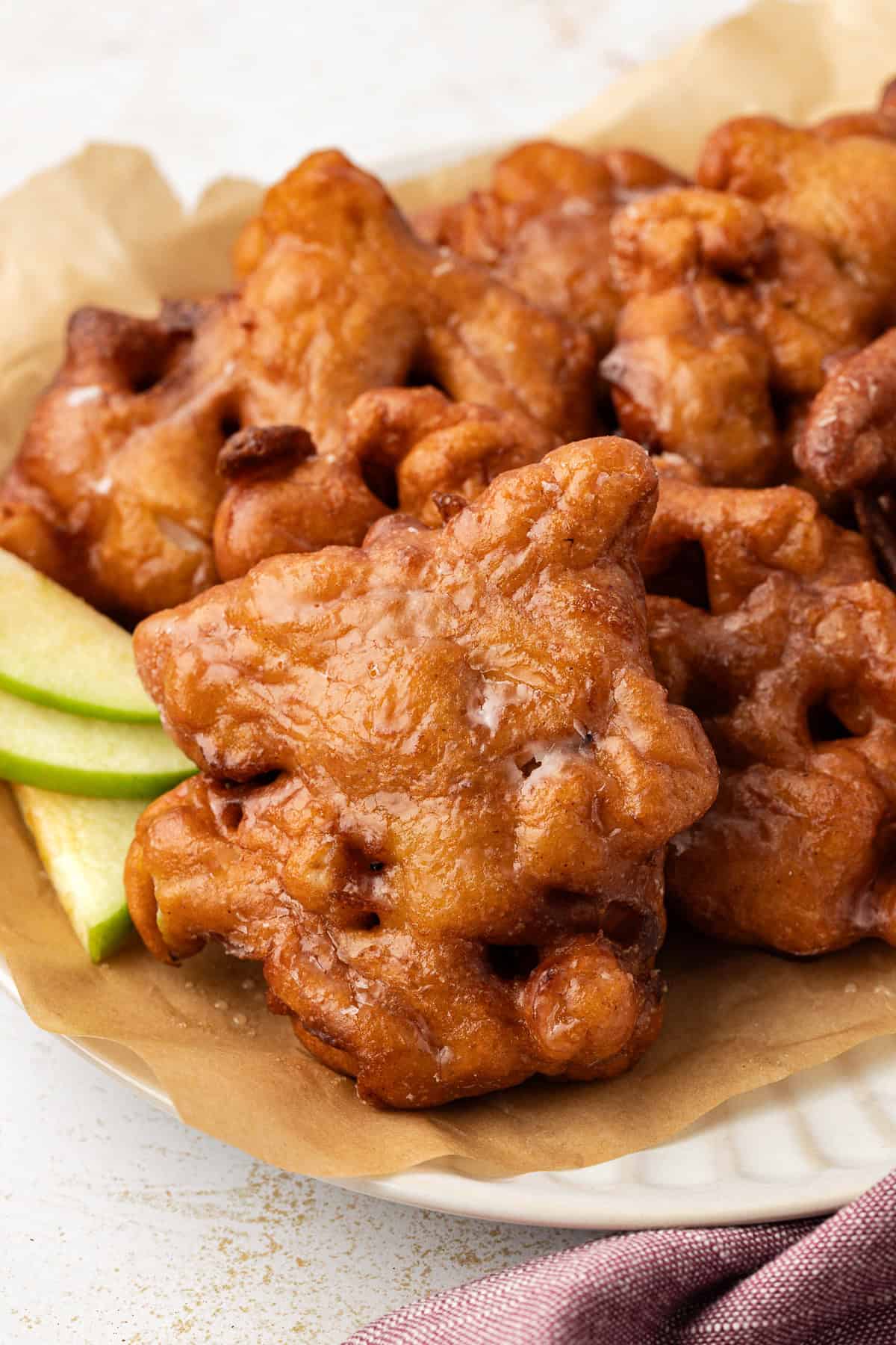 apple fritters piled on a plate covered in brown paper with three fresh apple slices