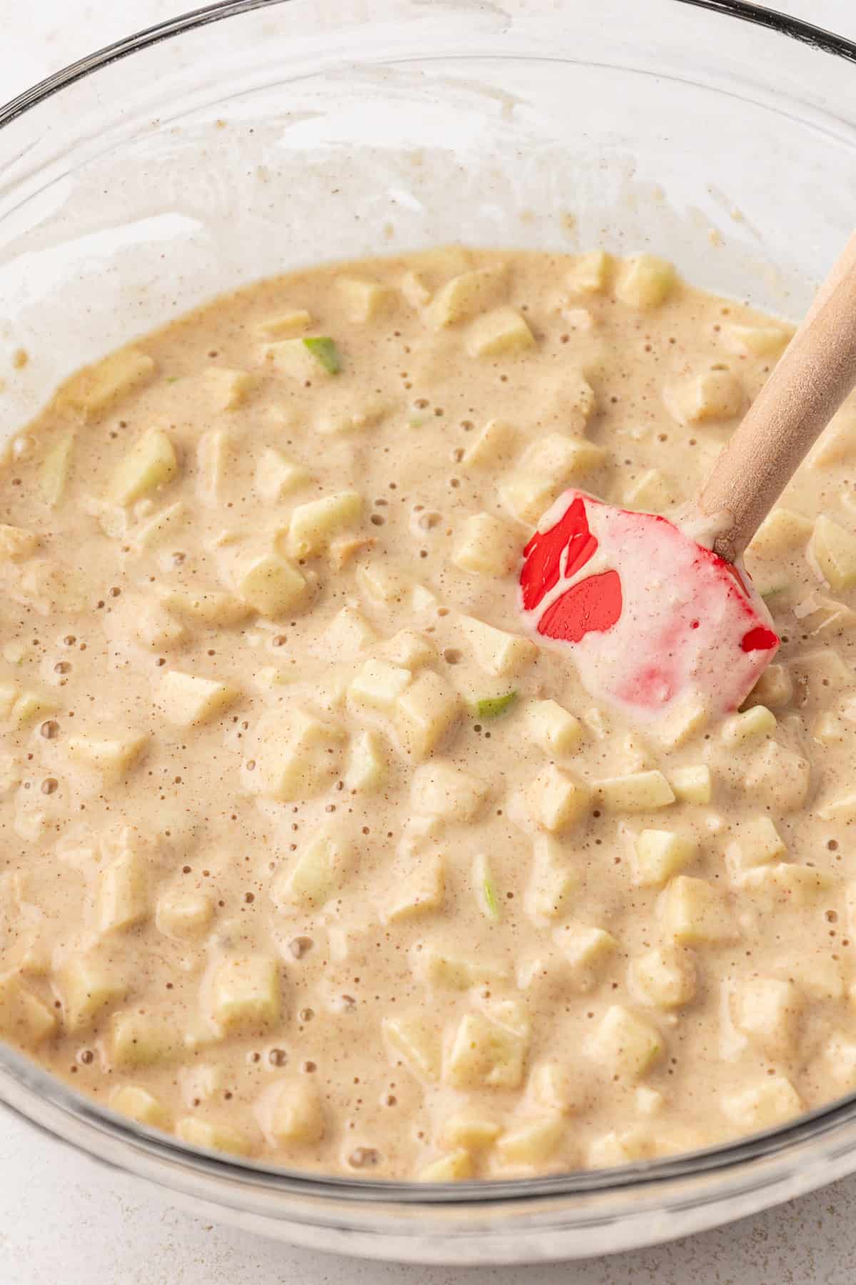 apple fritter batter in a clear glass bowl with a red spatula