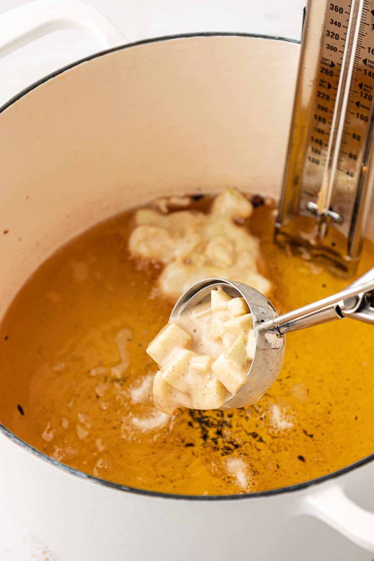 a cookie scoop dropping apple fritter batter into a pot of hot oil with a thermometer in it