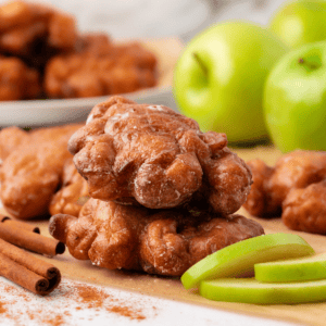 two apple fritters stacked on brown paper, surrounded by green apple slices, cinnamon sticks, and more fritters and whole green apples in the background