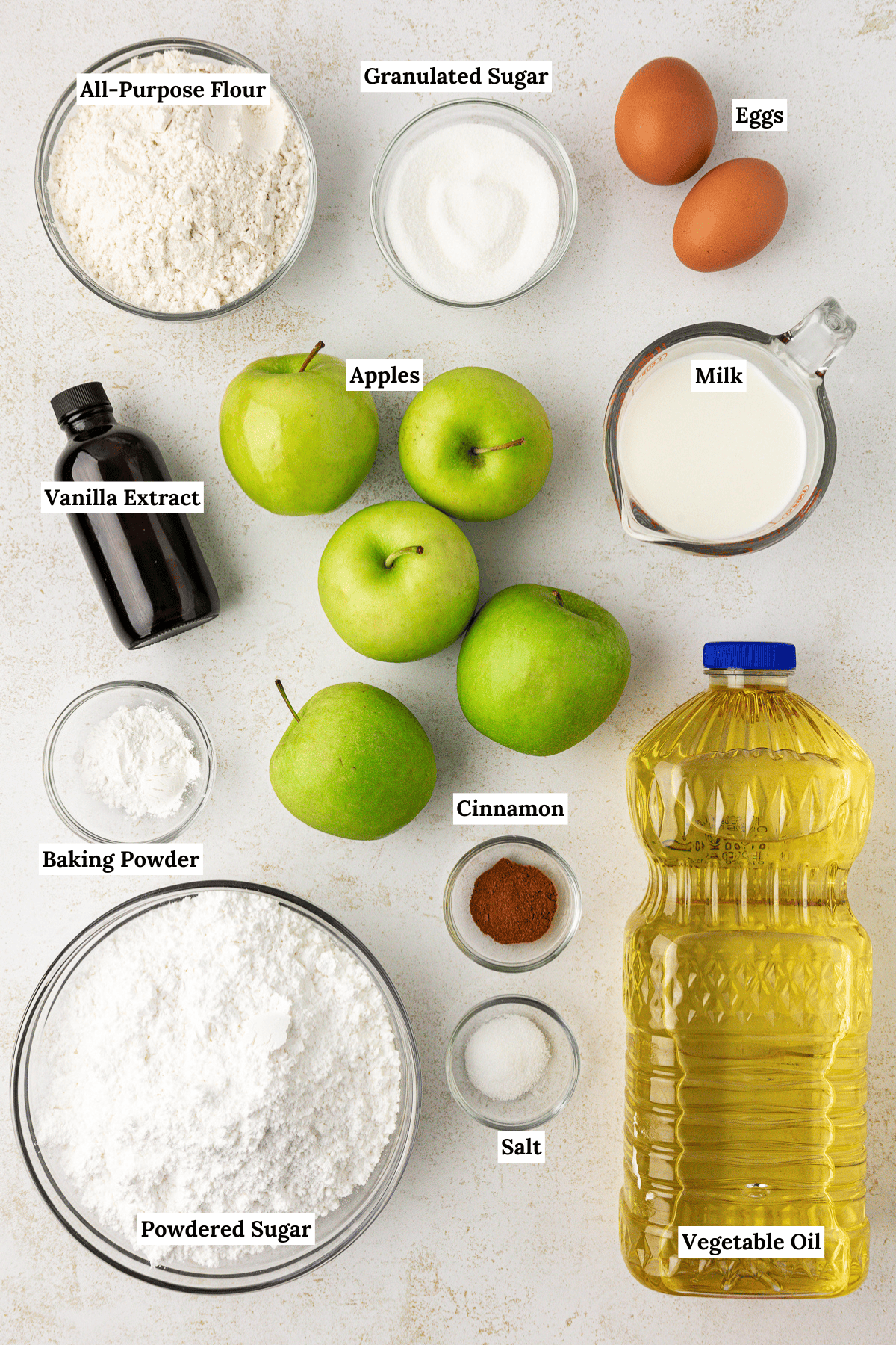 over head view of the ingredients for apple fritters, including all-purpose flour, granulated sugar, eggs, vanilla extract, apples, milk, baking powder, cinnamon, salt, vegetable oil, and powdered sugar