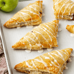 apple turnovers drizzled with glaze in a row on a baking sheet lined with parchment paper, surrounded by two whole green apples and cinnamon sticks