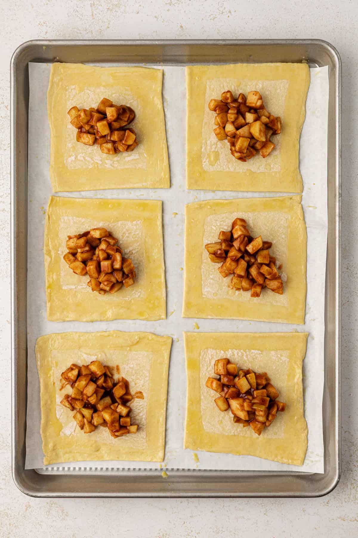 6 squares of puff pastry with a dollop of apple filling in the middle, in two rows on a baking sheet lined with parchment paper