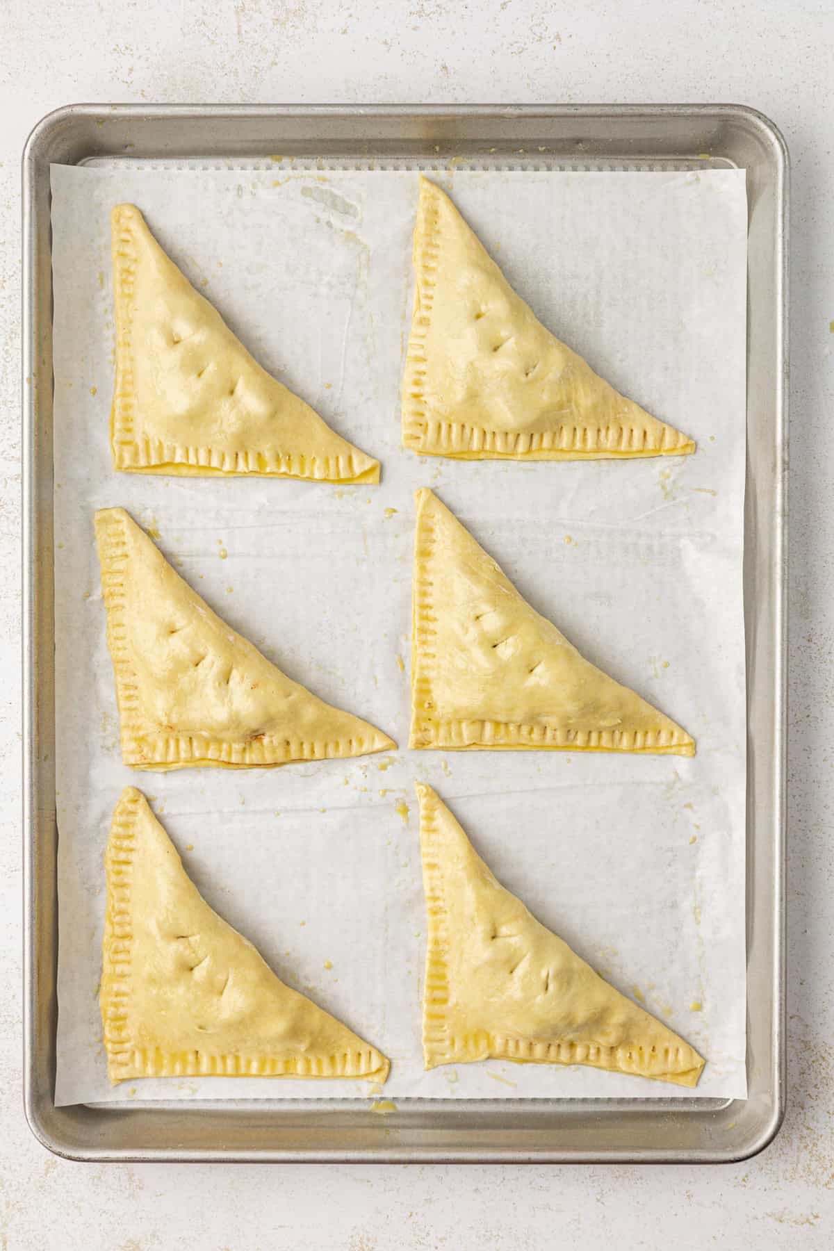 six apple turnovers in two rows on a baking sheet lined with parchment paper, ready to bake