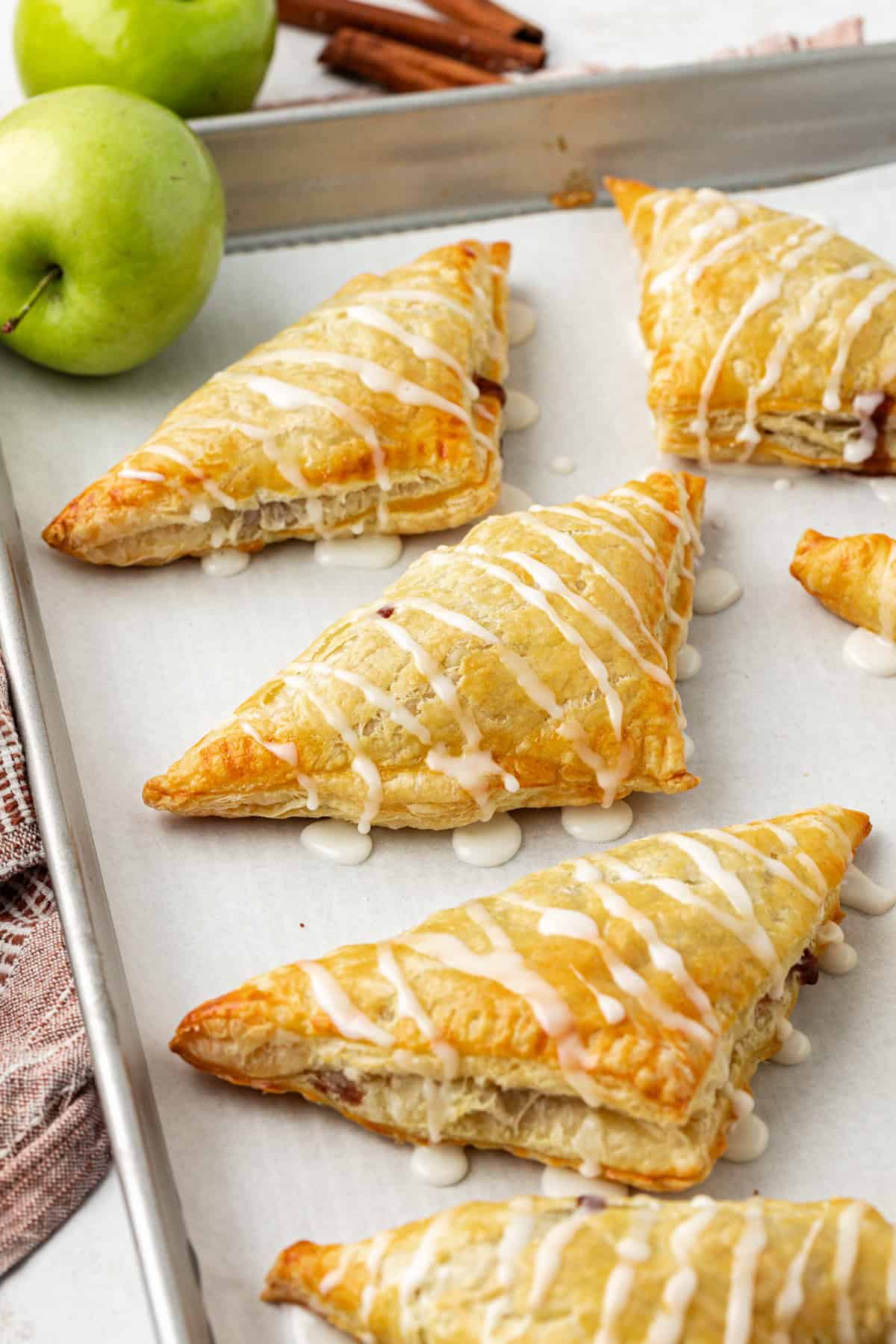 apple turnovers drizzled with glaze in a row on a baking sheet lined with parchment paper, surrounded by two whole green apples and cinnamon sticks