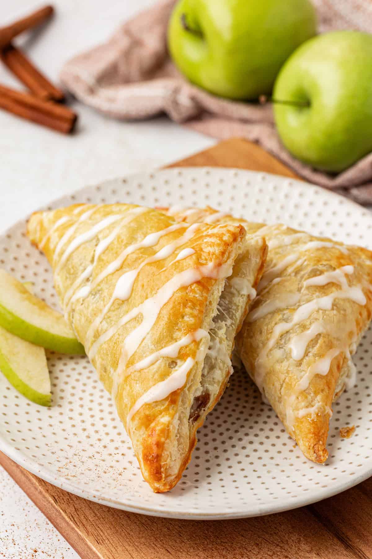 two apple turnovers on a small white plate with two green apple slices, two whole green apples and cinnamon sticks in the background