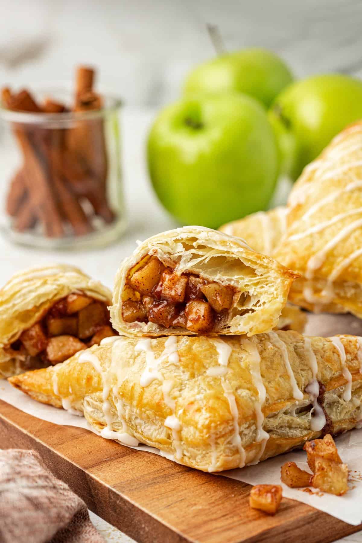 two halves of an apple turnover, showing the inside with apple filling, with half leaning on top of a whole turnover and more turnovers, whole green apples and cinnamon sticks in the background