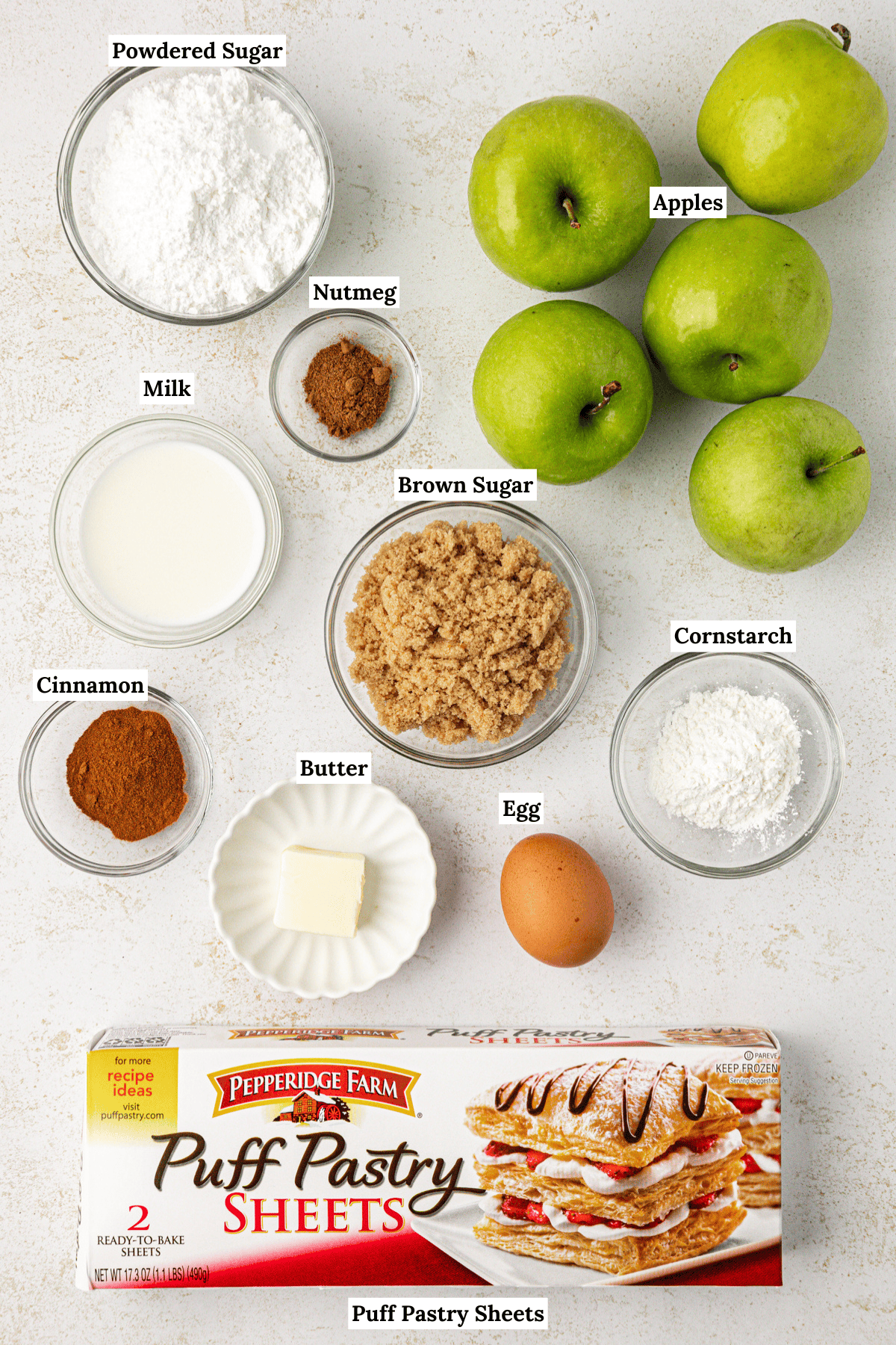 overhead view of the ingredients for apple turnovers including powdered sugar, nutmeg, apples, milk, brown sugar, cinnamon, butter, egg, cornstarch, and puff pastry