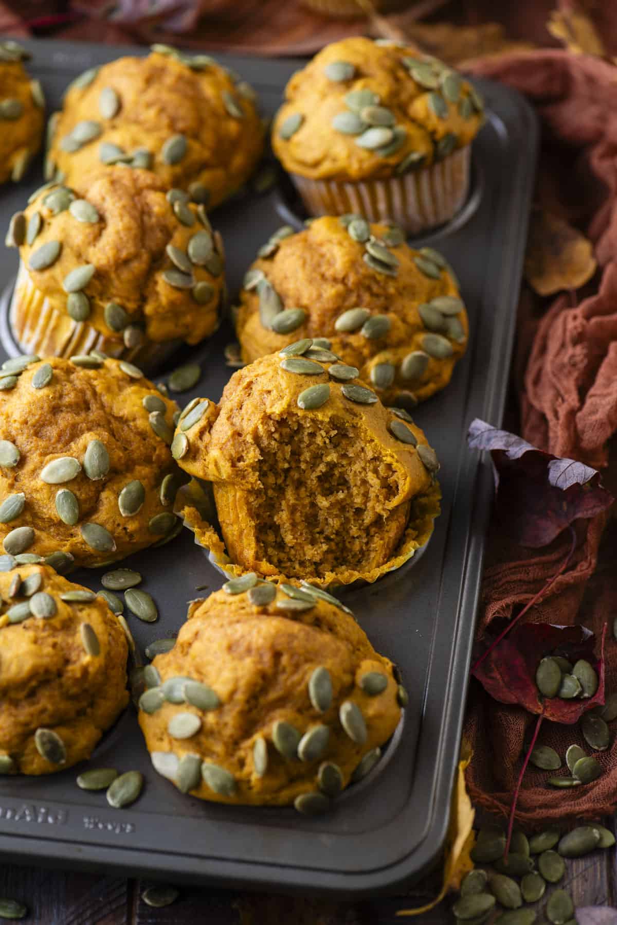 pumpkin muffins topped with pumpkin seeds in a muffin pan sitting on a dark orange towel, surrounded by pumpkin seeds and fall leaves