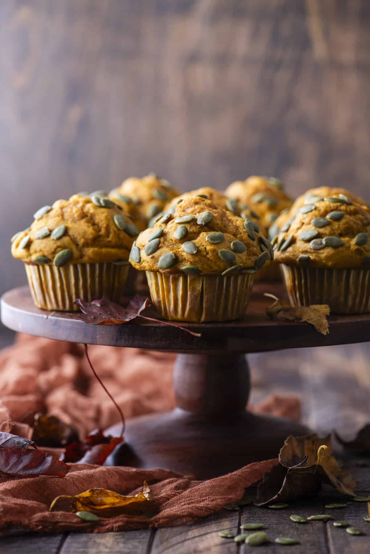 pumpkin muffins topped with pumpkin seeds on a wood cake stand surrounded by pumpkin seeds and fall leaves