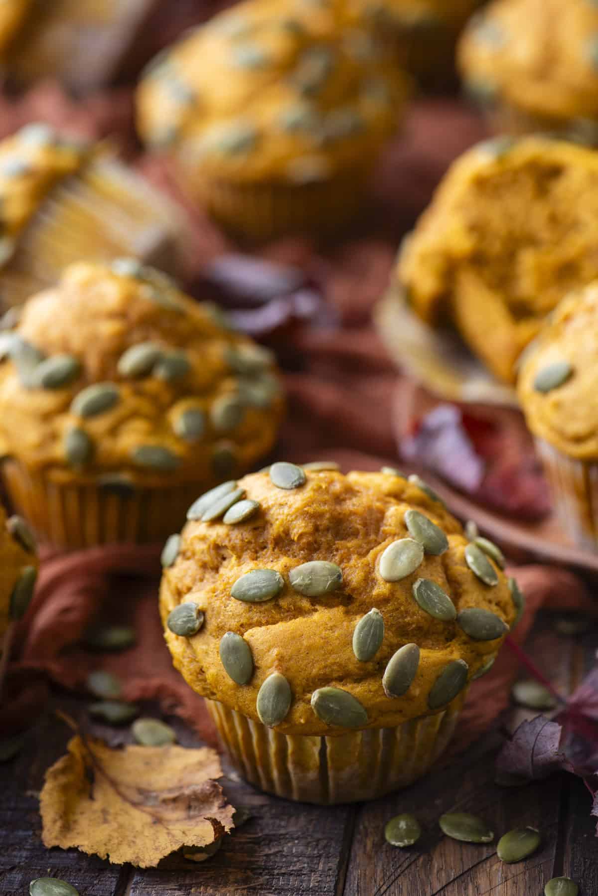 a healthy pumpkin muffin topped with pumpkin seeds surrounded by pumpkin seeds, fall leaves and more muffins scattered in the background on a dark orange towel