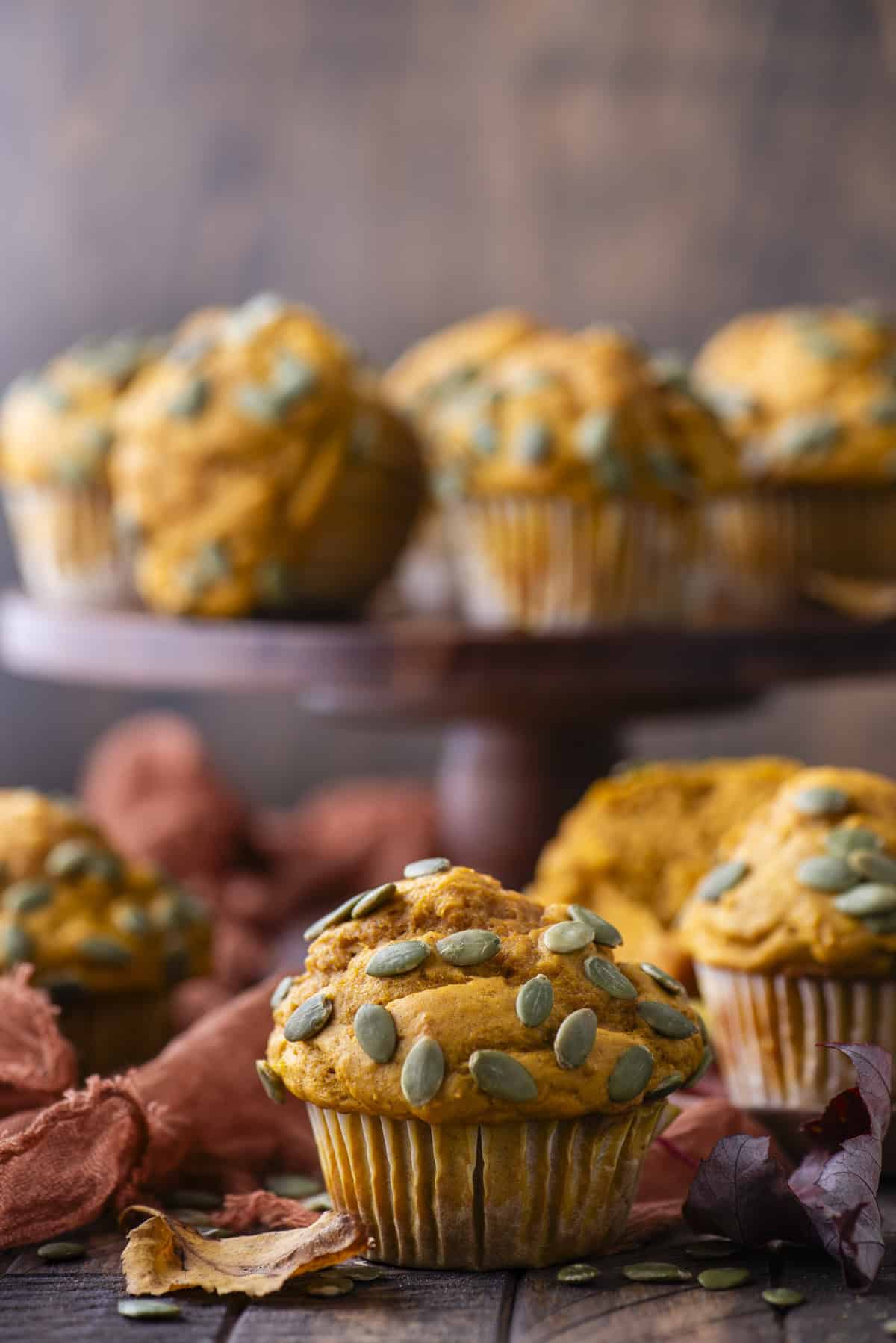 a pumpkin muffin surrounded by pumpkin seeds, fall leaves and a dark orange towel with more pumpkin muffins scattered in the background and on a wood cake stand