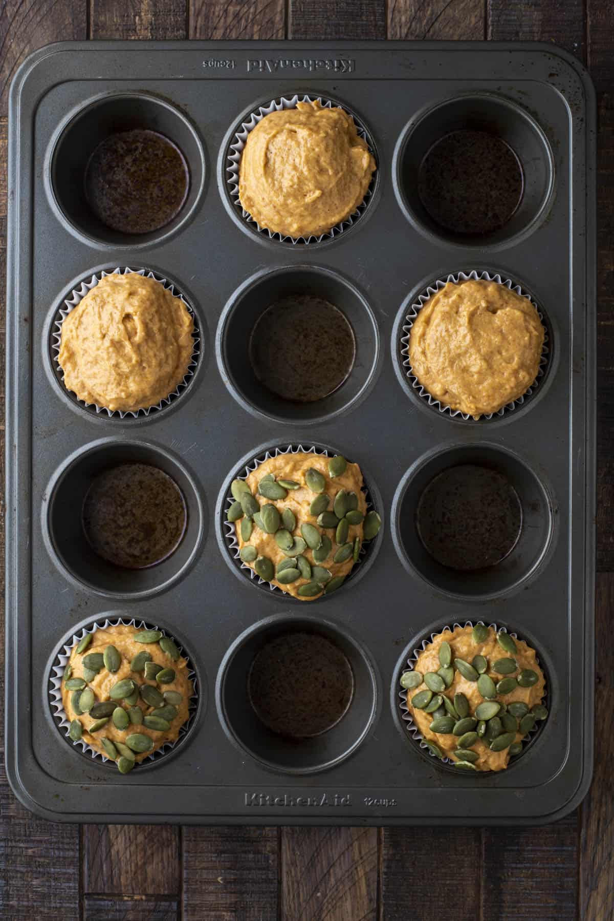 healthy pumpkin muffin batter in every other hole of a muffin pan with 3 topped with pumpkin seeds and 3 without