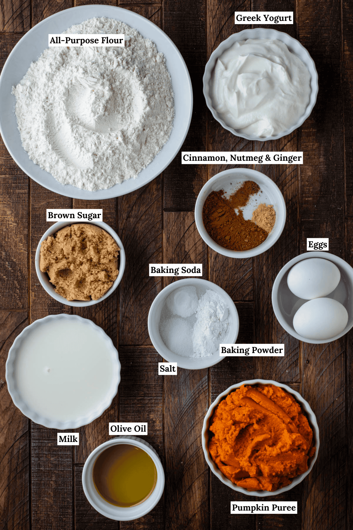 overhead view of the ingredients for healthy pumpkin muffins, including all-purpose flour, greek yogurt, brown sugar, cinnamon, nutmeg, ginger, milk, baking soda, salt, baking powder, eggs, olive oil, pumpkin puree