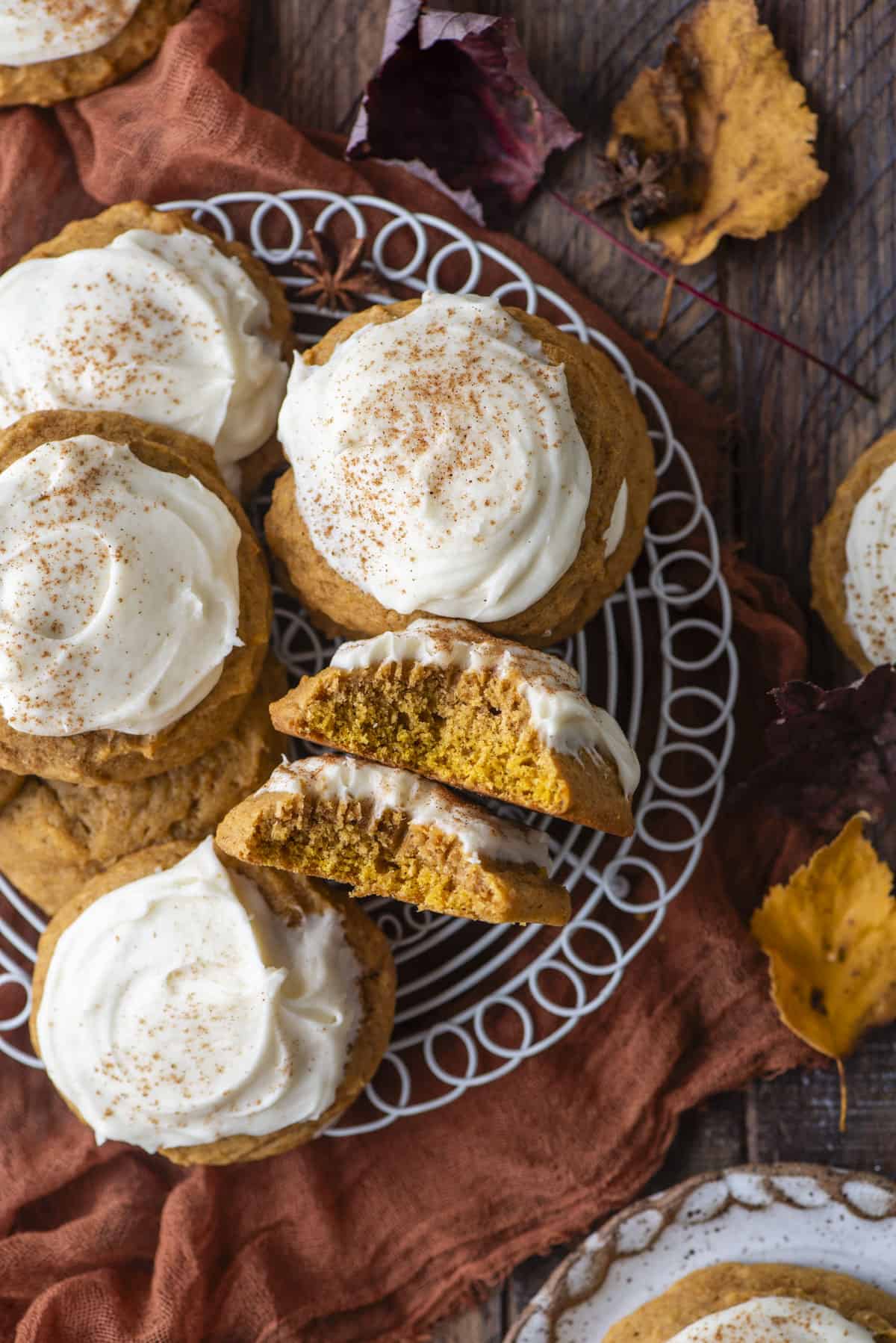 pumpkin cookies with cream cheese frosting on a white wire rack, with one cookie split in half and on its side showing the layers of cookie and frosting, surrounded by fall leaves