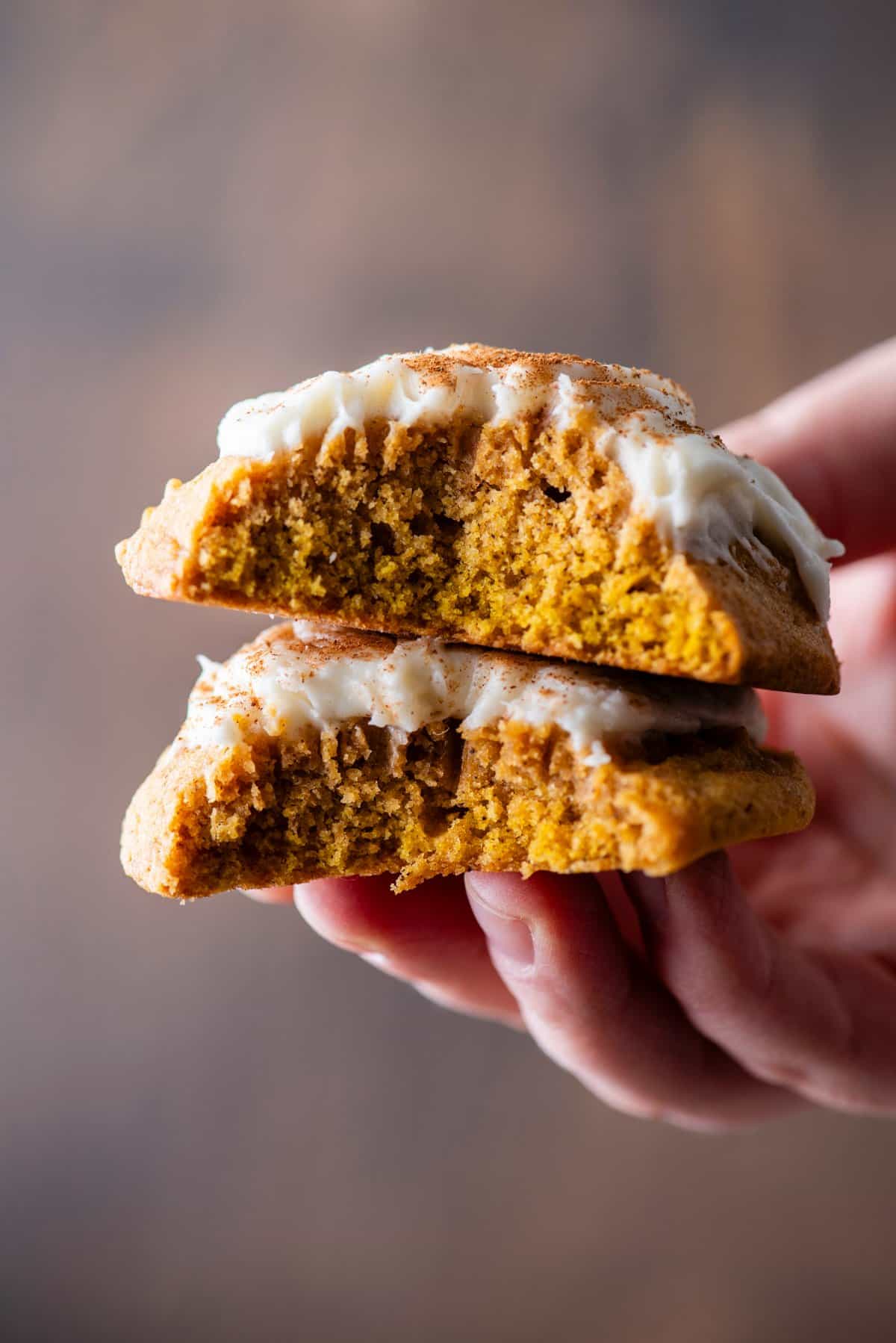 a hand holding a pumpkin cookie split in half and stacked together, showing the layers of cookie and cream cheese frosting