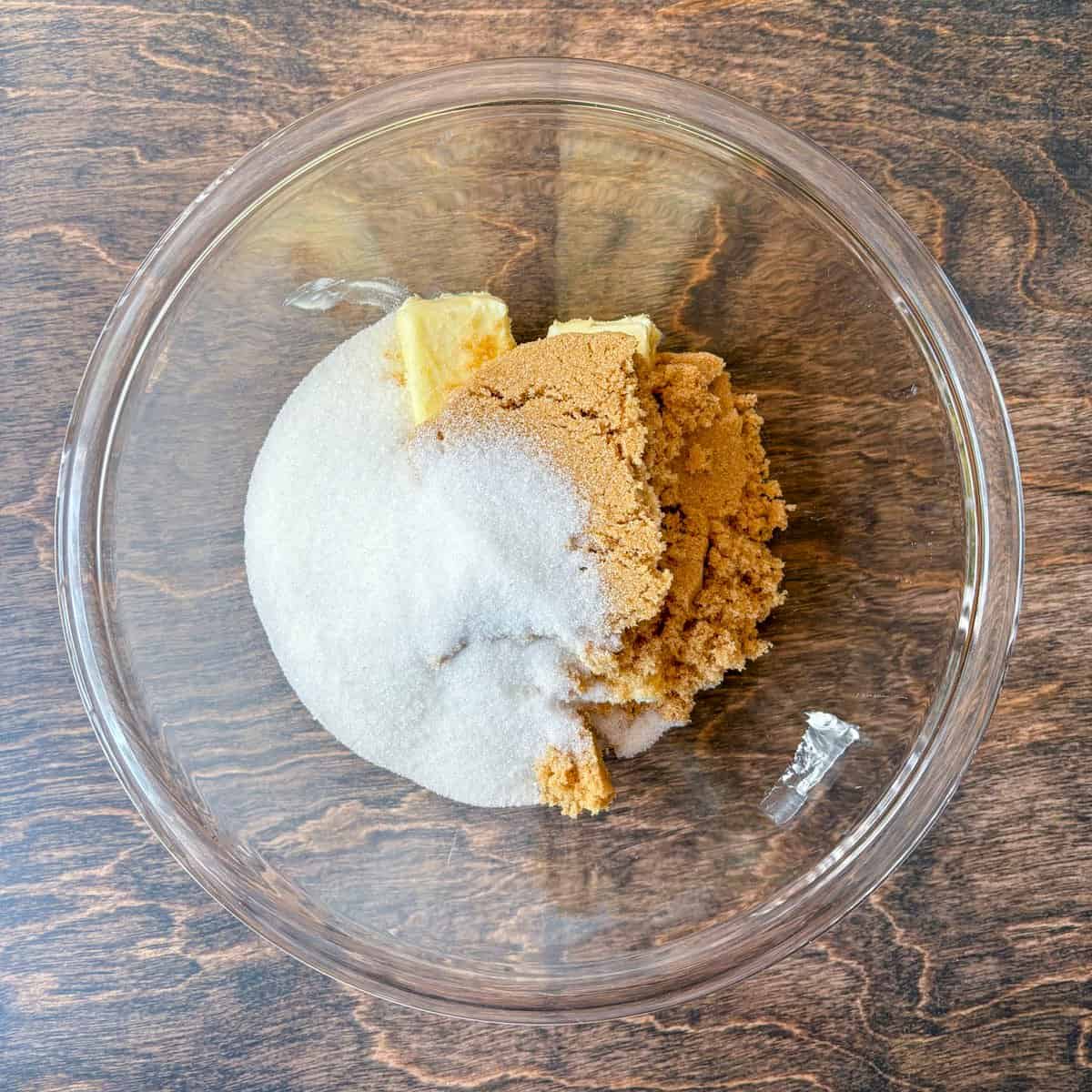 over head view of a clear glass bowl on a wood surface, with granulated sugar, brown sugar and butter in it