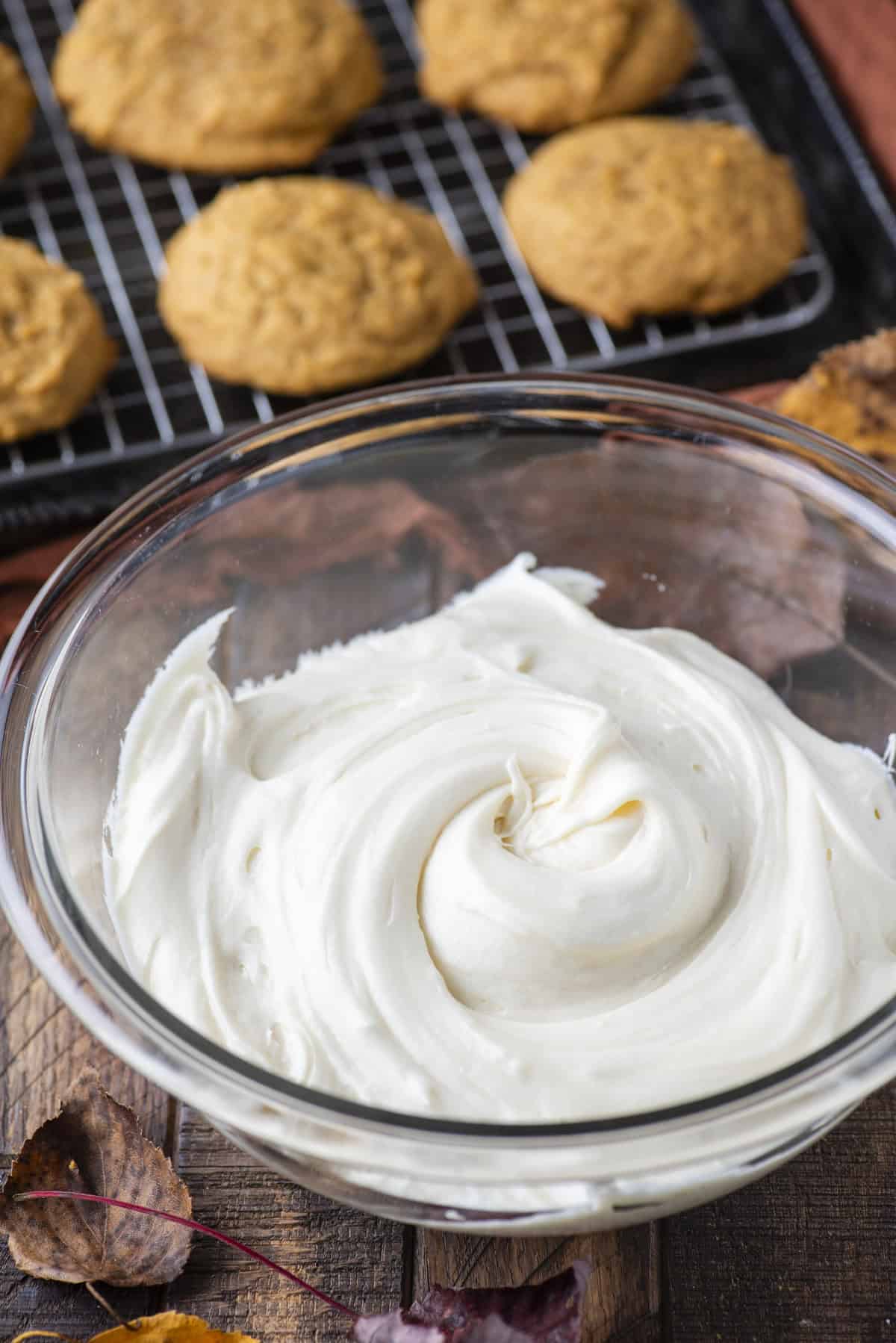 cream cheese frosting in a clear glass bowl surrounded by fall leaves beside a wire rack with pumpkin cookies in rows on it