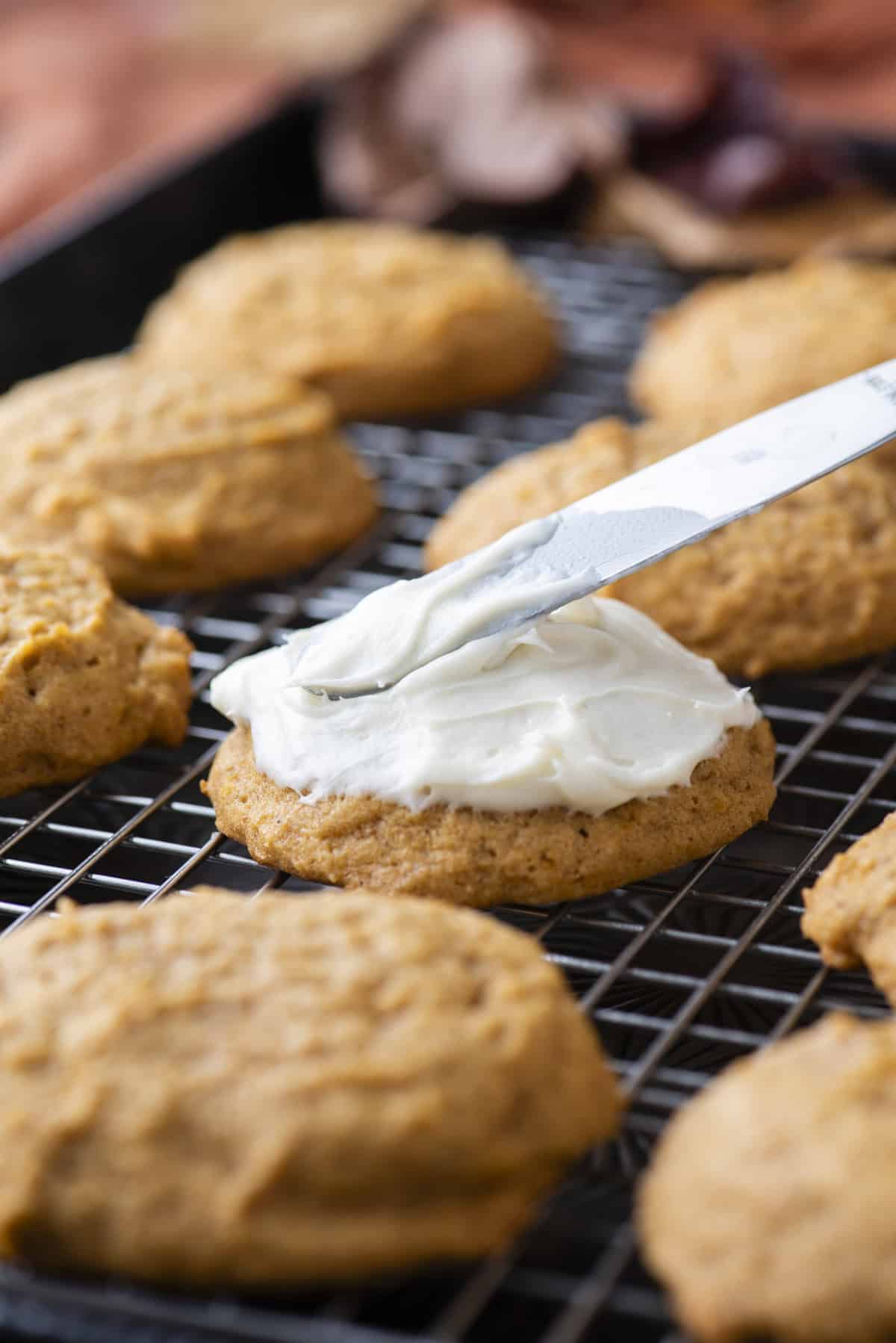 pumpkin cookies on a wire rack with one being topped with cream cheese frosting with a butter knife