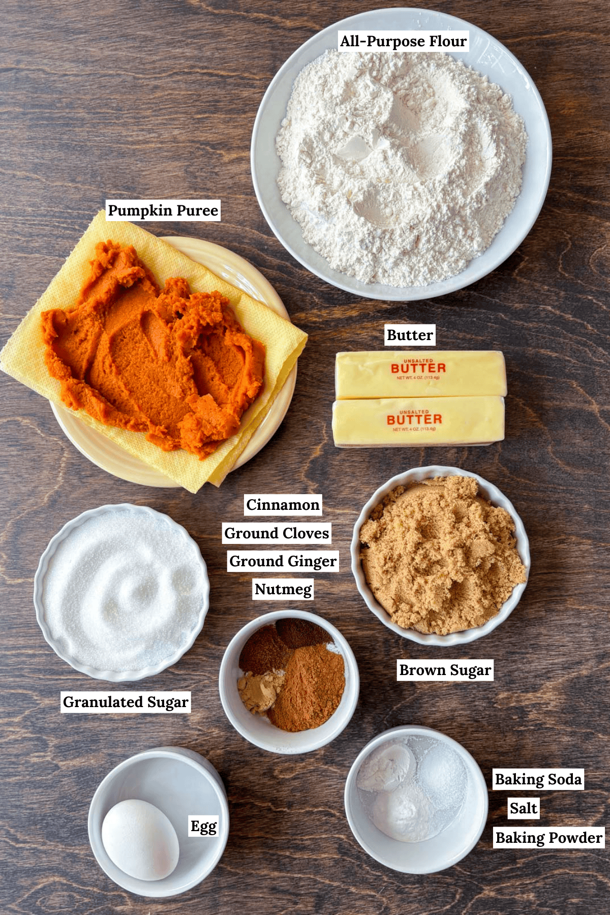 over head view of the ingredients for pumpkin cookies including all-purpose flour, pumpkin puree, butter, granulated sugar, cinnamon, ground cloves, ground ginger, nutmeg, brown sugar, egg, baking soda, salt and baking powder