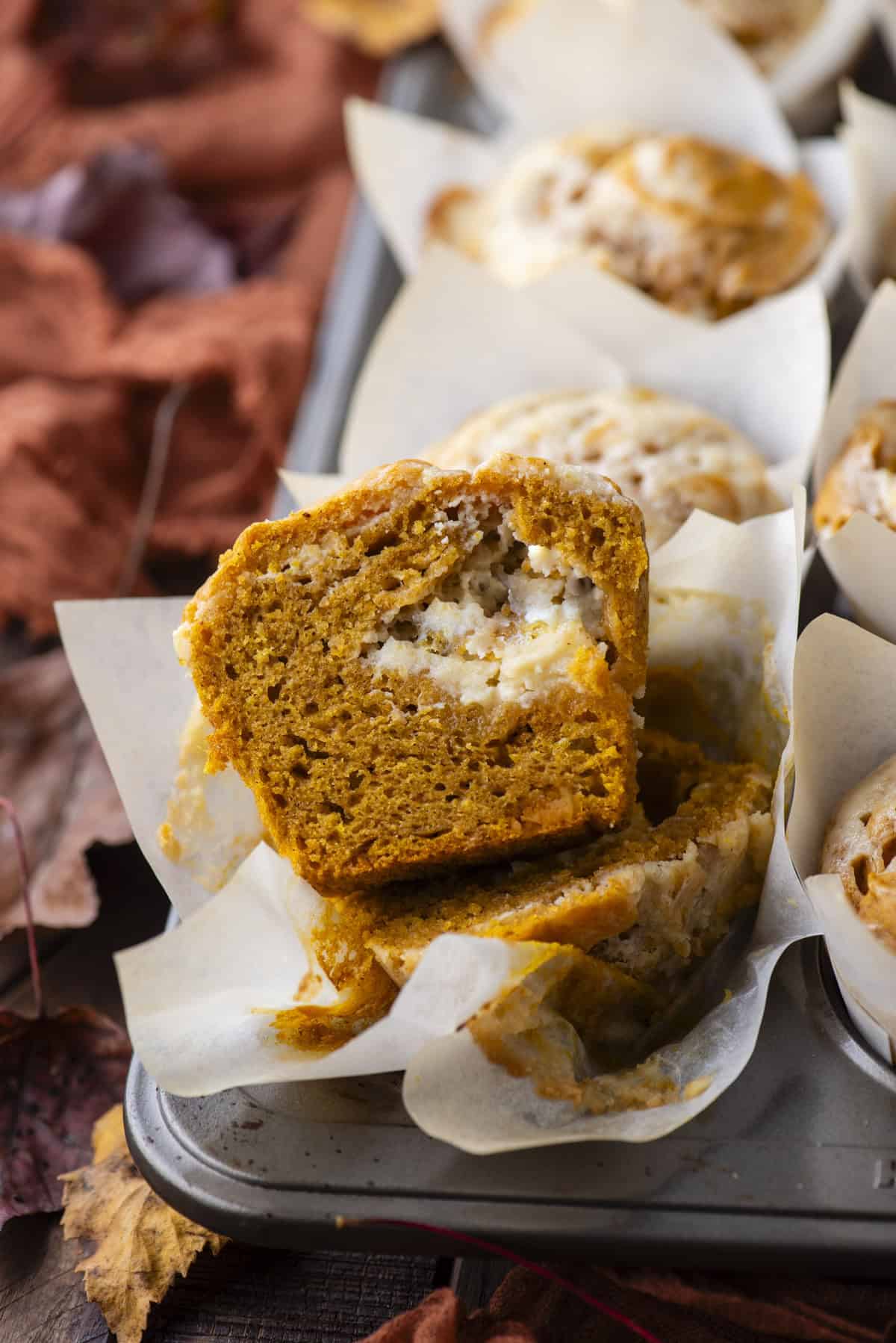 a pumpkin cream cheese muffin cut in half exposing the cream cheese layer inside, sitting in its muffin liner in a muffin pan full of muffins