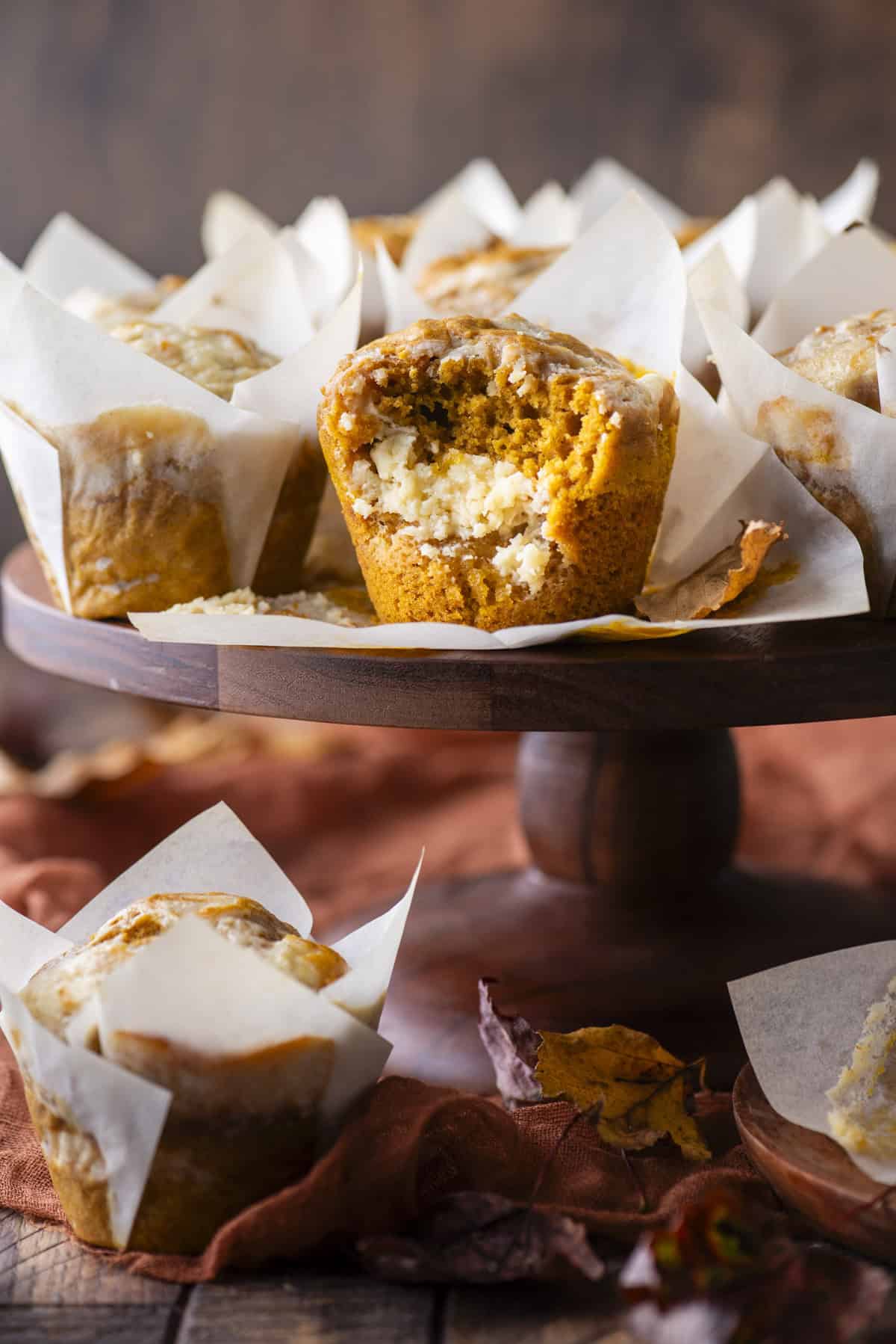 a pumpkin cream cheese muffin with a bite missing out of it, exposing the cream cheese layer inside, sitting on top of a wood cake stand with more muffins on top and below, and fall leaves scattered around