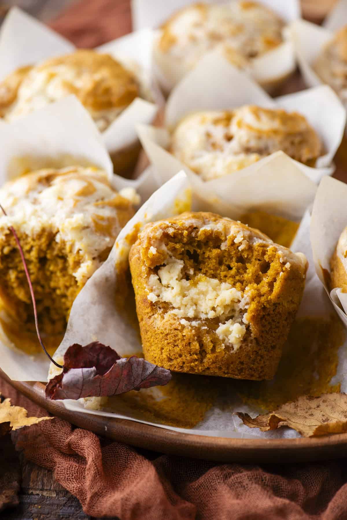 a pumpkin cream cheese muffin with a bite missing out of if, on a wood platter full of muffins surrounded by fall leaves