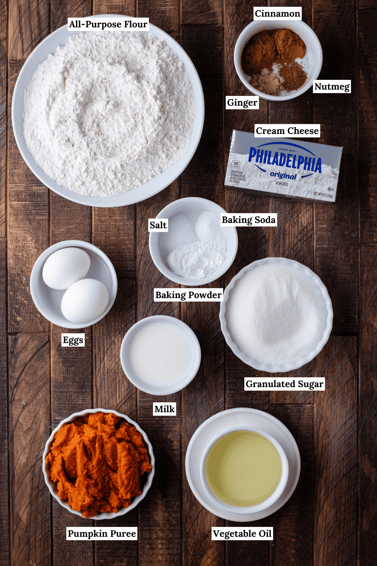 over head view of the ingredients for pumpkin cream cheese muffins including all-purpose flour, cinnamon, ginger, nutmeg, cream cheese, eggs, salt, baking soda, granulated sugar, milk, pumpkin puree, and vegetable oil