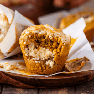 a pumpkin cream cheese muffin with a bite missing out, exposing the cream cheese layer inside, on a wood platter with more muffins and fall leaves