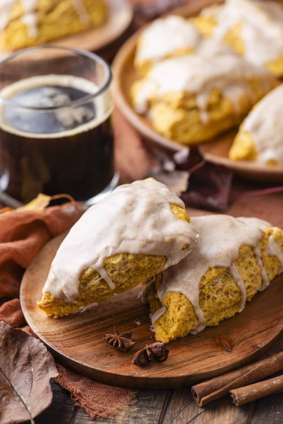 two glazed pumpkin scones on a wood platter with star anise, fall leaves and cinnamon sticks scattered around and a cup of black coffee and more scones in the background