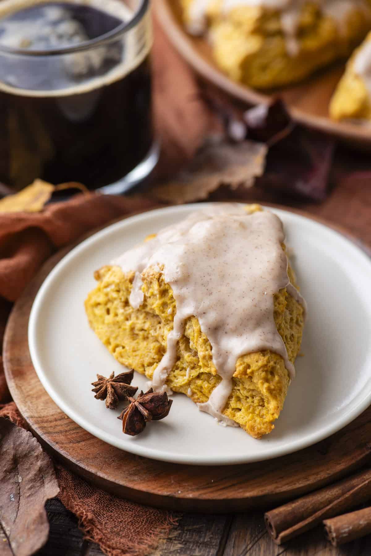 a glazed pumpkin scone on a small white plate on top of a wood platter, with star anise beside it, a cup of coffee in the background and fall leaves and cinnamon sticks scattered around