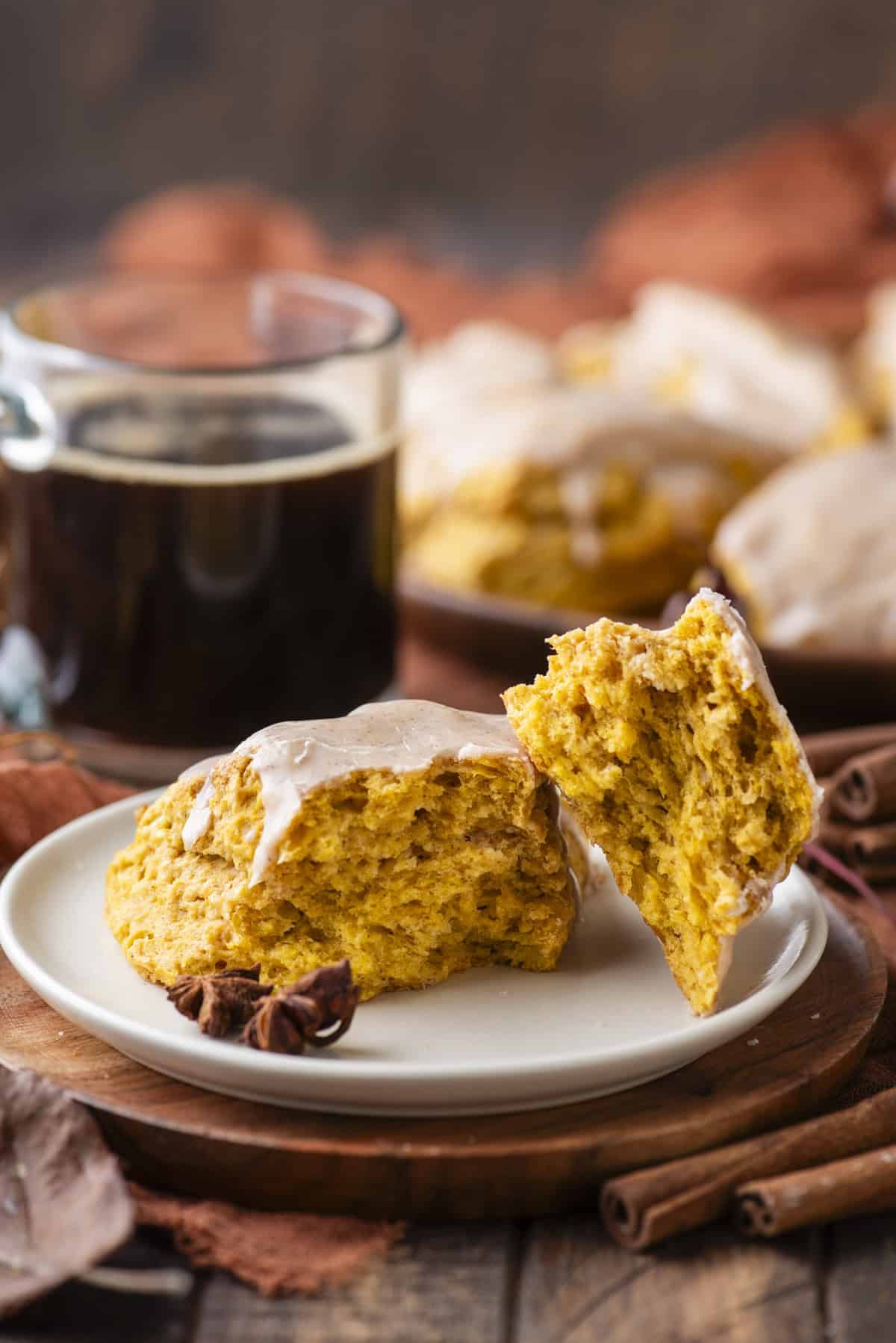 a pumpkin scone broken in half with one half leaning on the other on a small white plate on top of a wood platter, with a cup of coffee and more scones in the background and fall leaves, cinnamon sticks and star anise scattered around