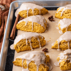 a baking sheet lined with a silicone mat with two rows of glazed pumpkin scones on it, fall leaves, cinnamon sticks and star anise scattered around