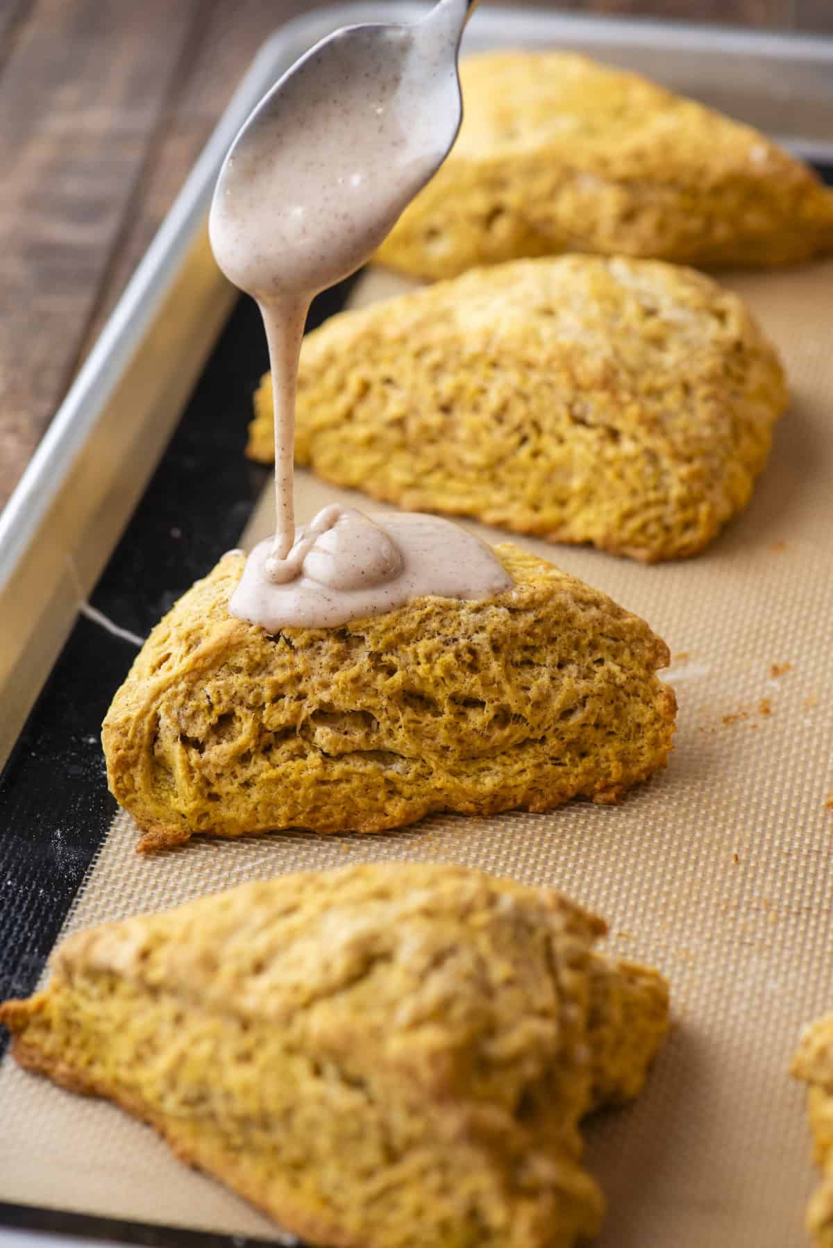 a glaze being drizzled over a pumpkin scone on a baking sheet with rows of scones