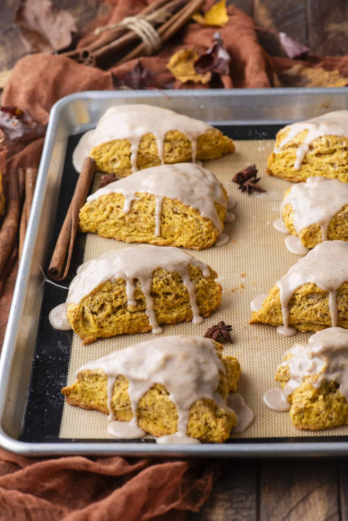 a baking sheet lined with a silicone mat with two rows of glazed pumpkin scones on it, fall leaves, cinnamon sticks and star anise scattered around