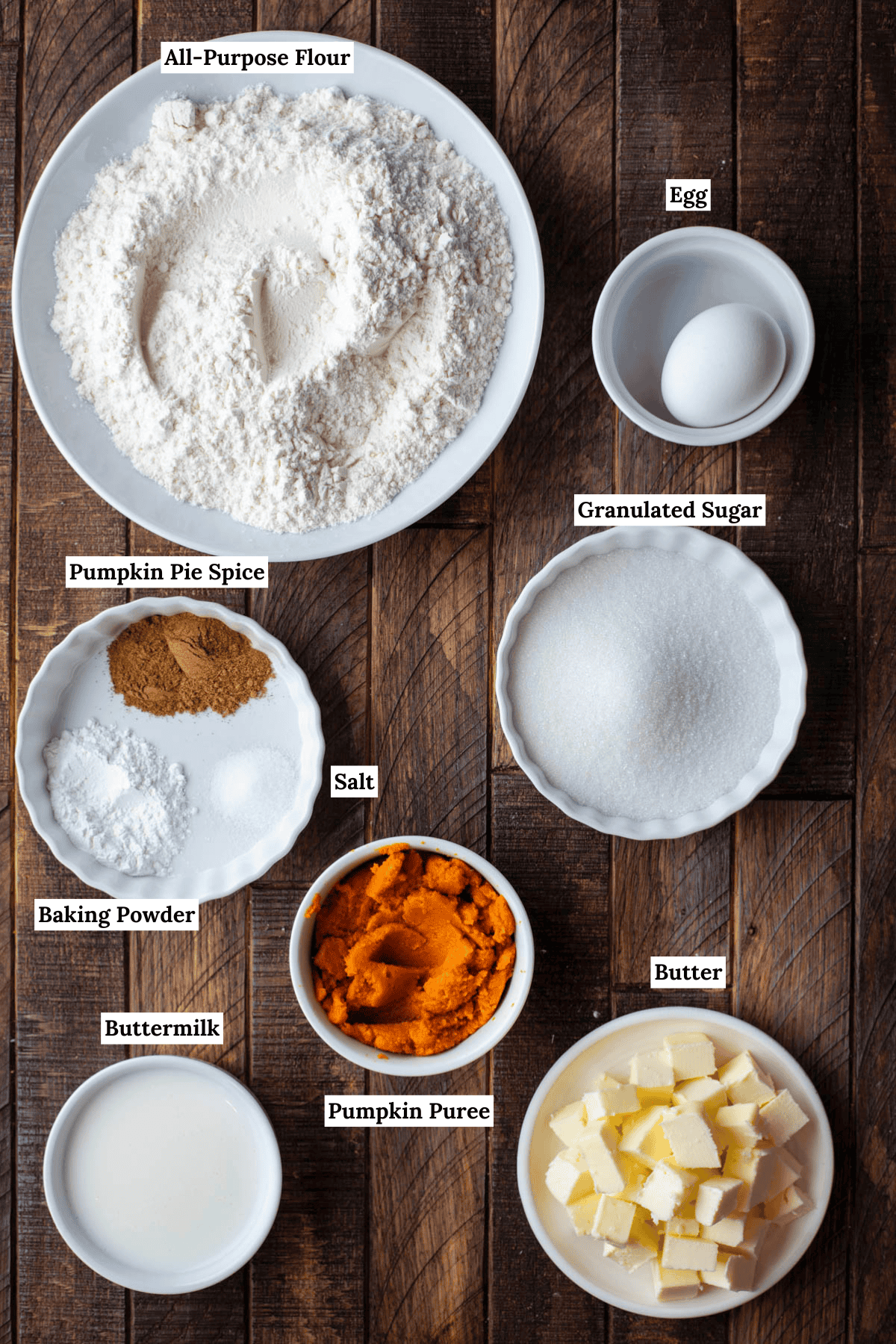 over head view of the ingredients for pumpkin scones including all-purpose flour, egg, pumpkin pie spice, baking powder, salt, granulated sugar, buttermilk, pumpkin puree and cubed cold butter