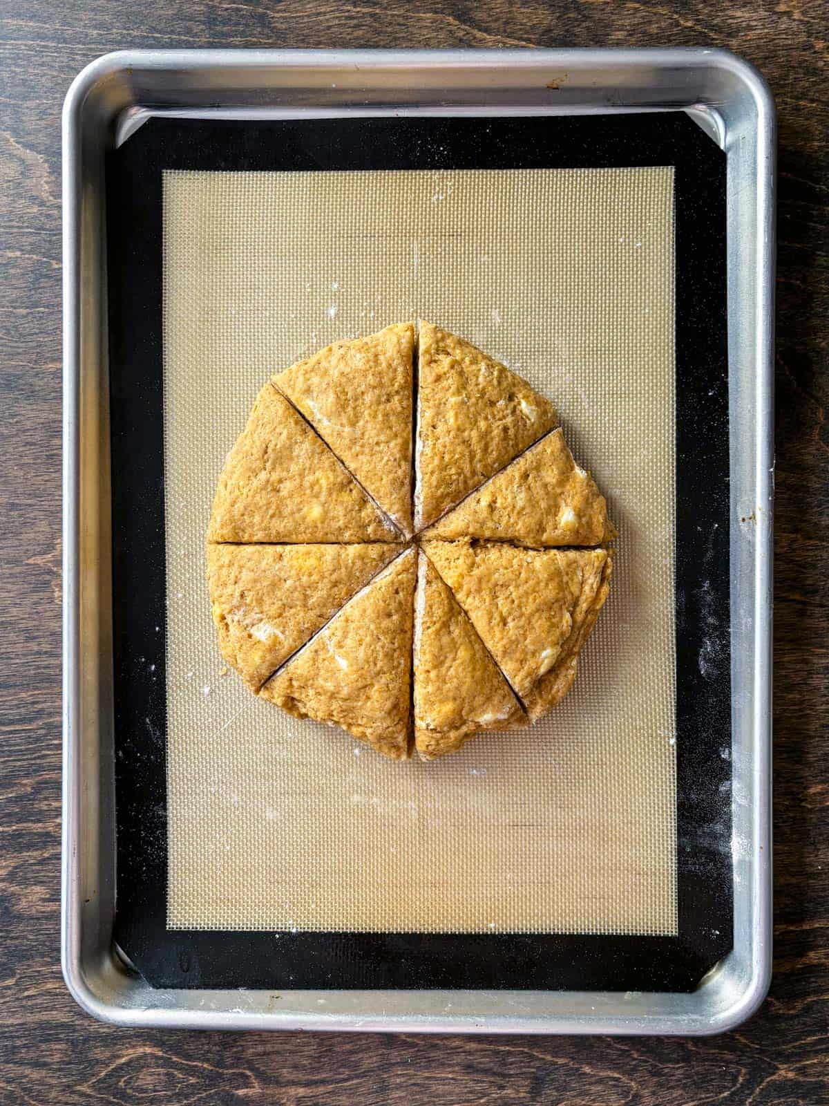 over head view of pumpin scone dough in a circle, sliced in triangles on a baking sheet lined with a silicone mat