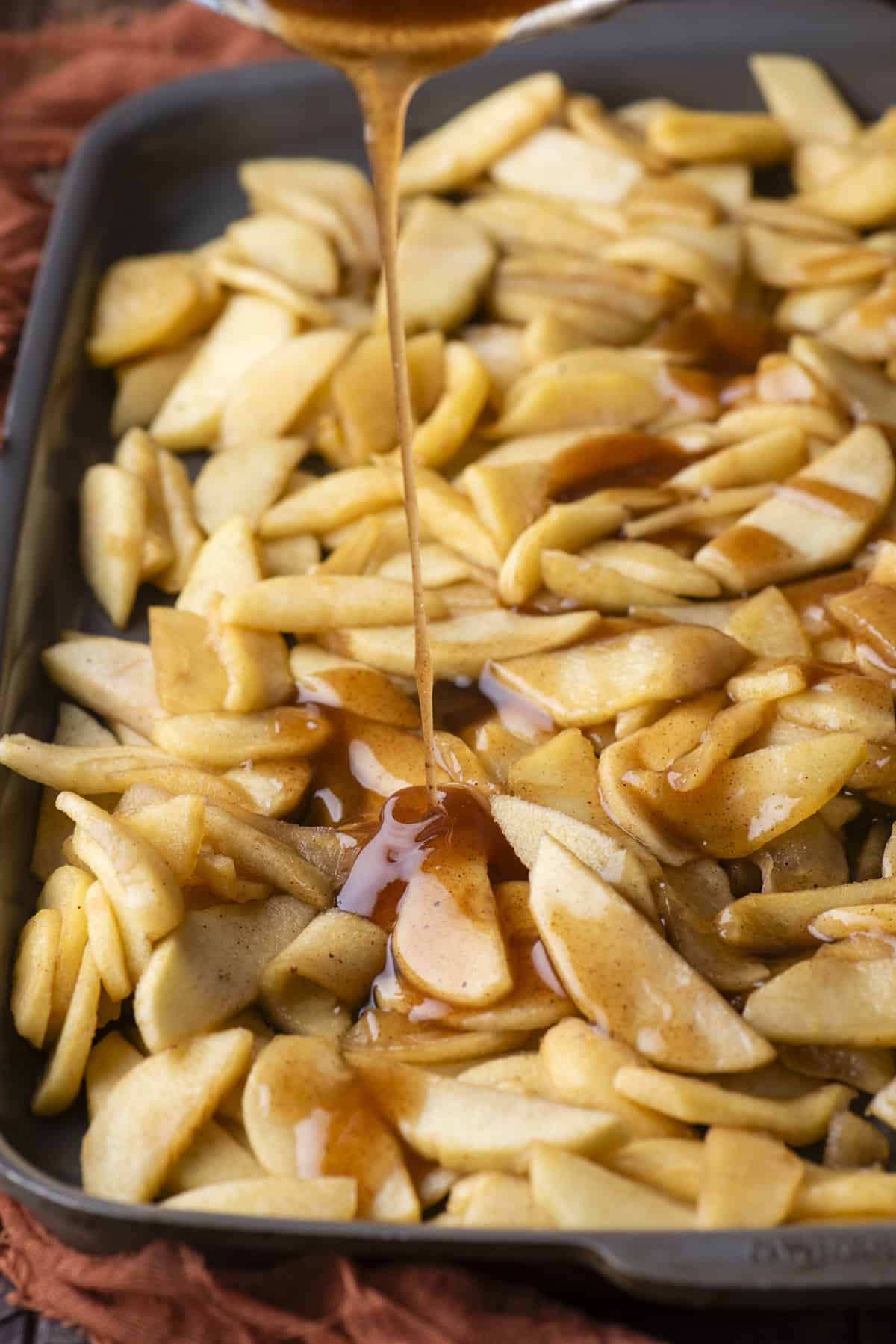 cooked apple slices spread out on a baking sheet with a syrup being poured over top of them