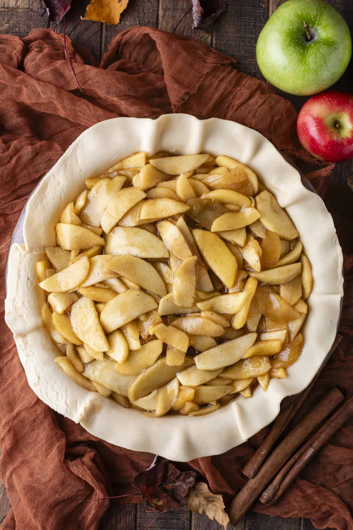 over head view of pie dish covered in uncooked pie crust and filled with apple filling, surrounded by green and red apples, cinnamon sticks and fall leaves