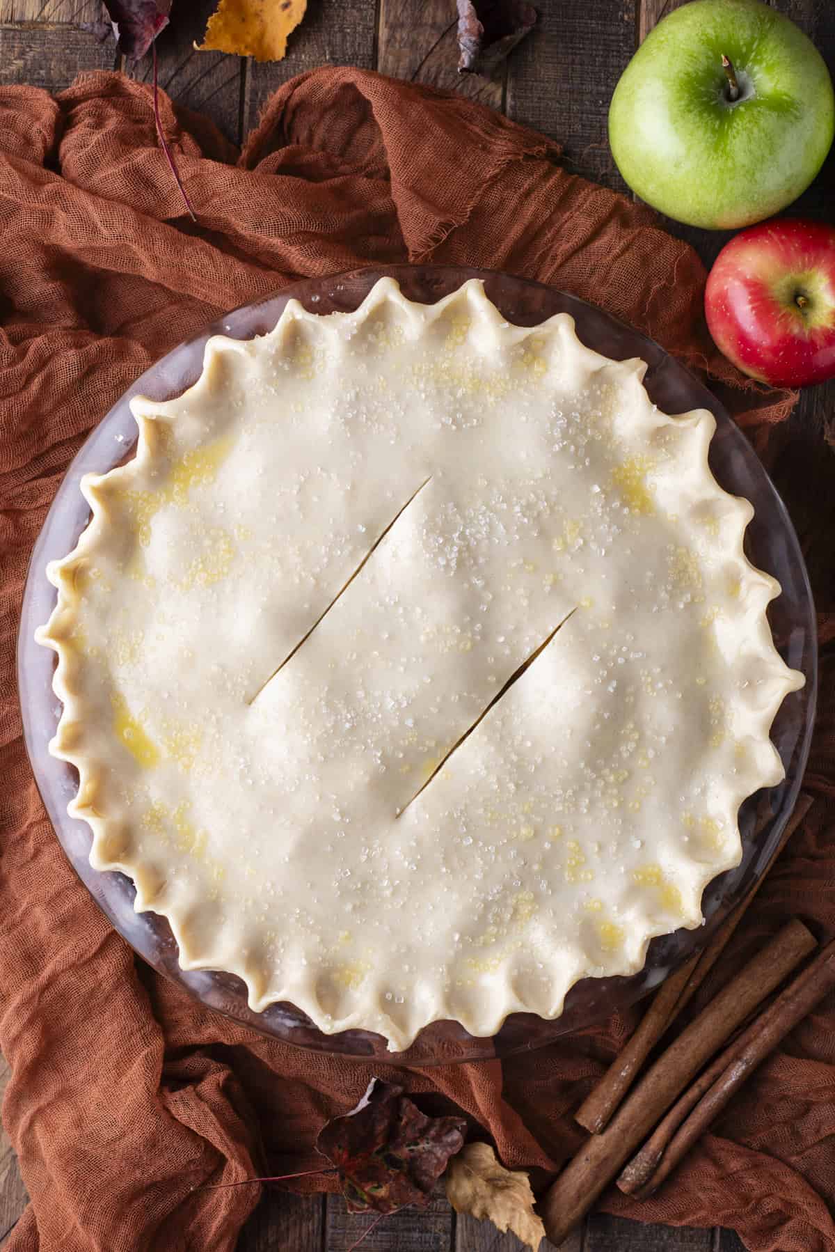 over head view of an unbaked apple pie surrounded by green and red apples, fall leaves and cinnamon sticks